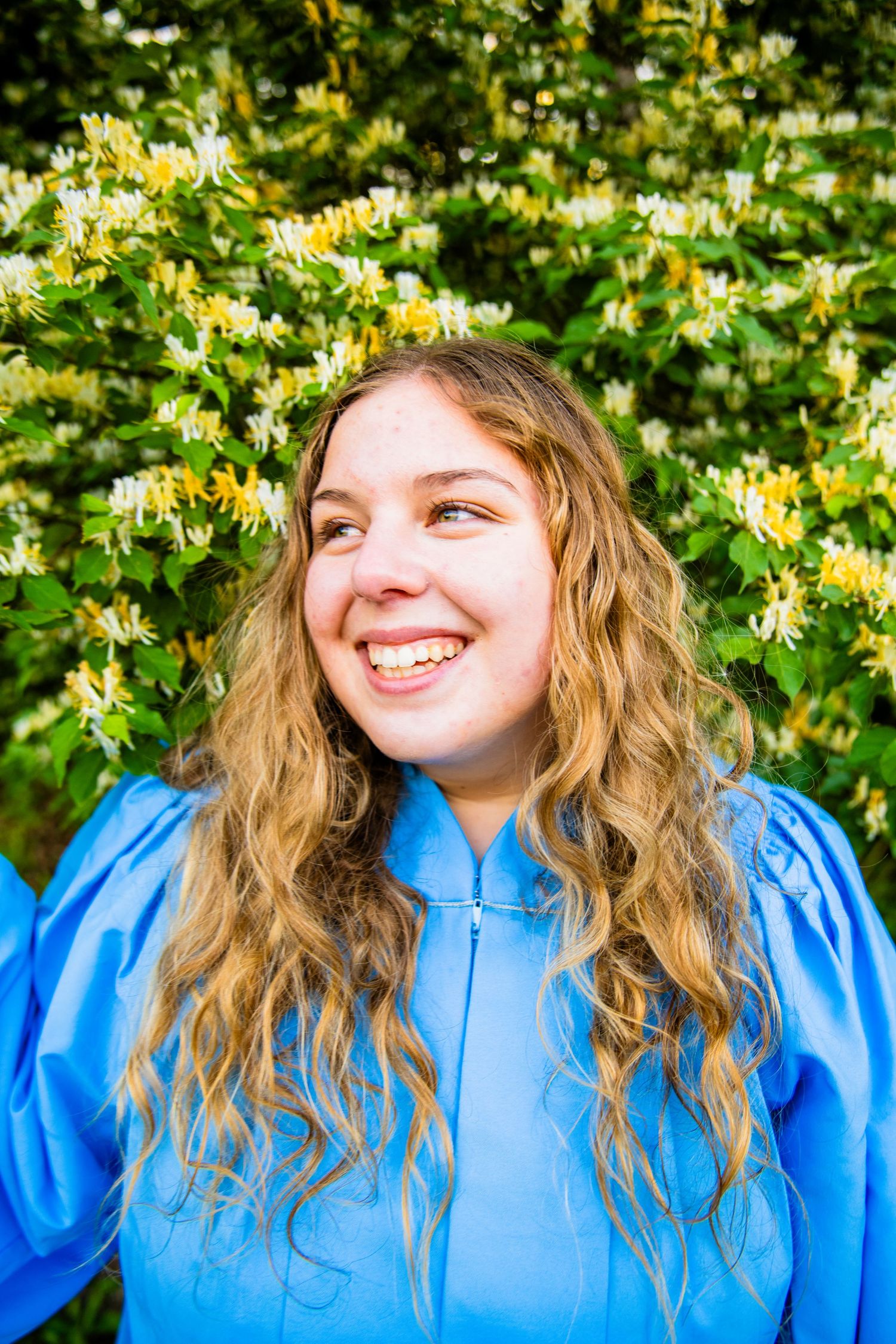 Smiling young woman with curly blonde hair wearing a blue graduation gown, posing in front of blooming honeysuckle bushes.