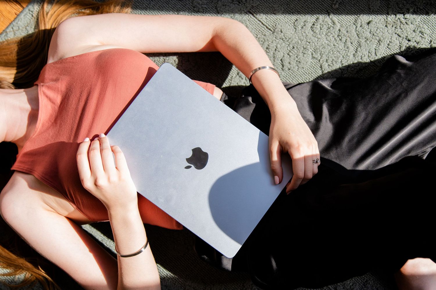 Person lying outdoors holding an Apple MacBook laptop over their face in sunlight.