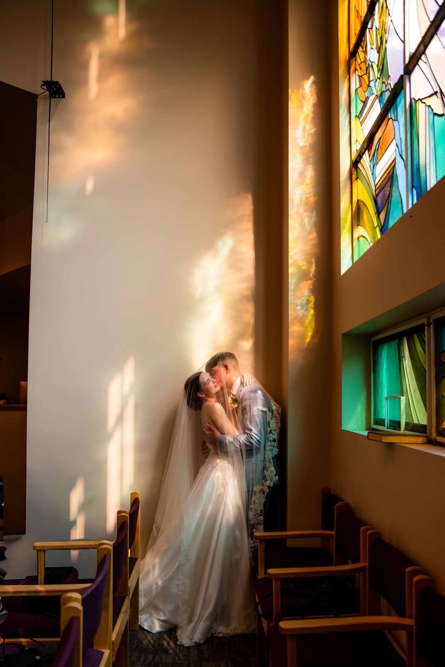 A bride and groom share a kiss in a church illuminated by colorful stained glass window light.