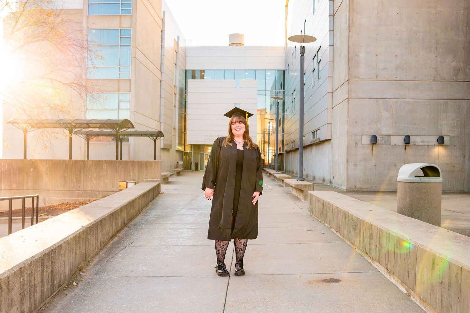 Graduate in cap and gown poses outdoors on campus walkway with golden sunlight and modern buildings.