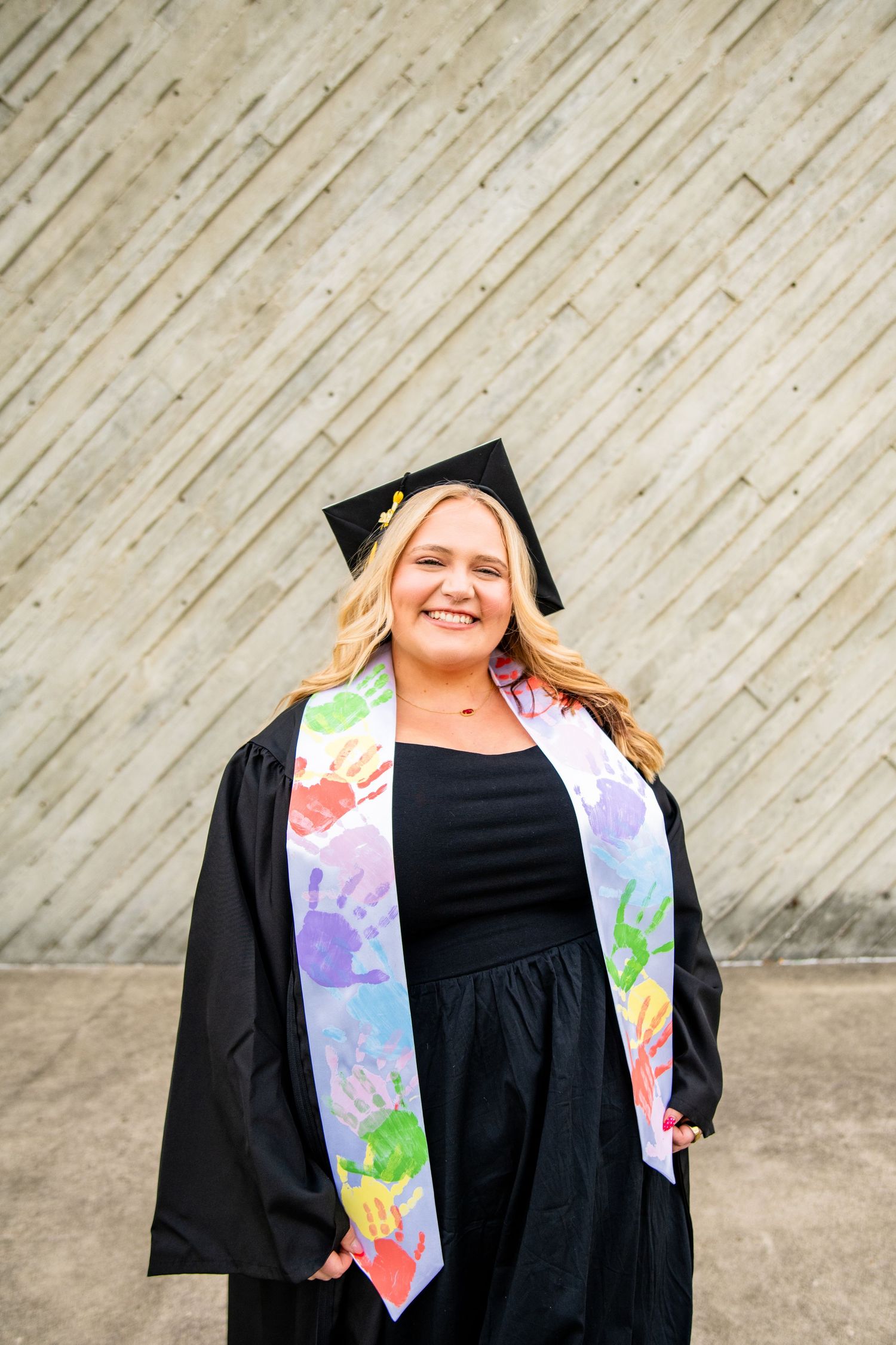 Smiling graduate in black cap and gown with colorful handprint stole posing outdoors.