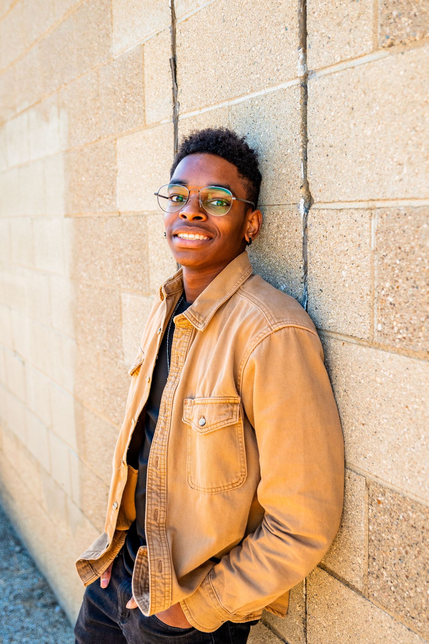 Young Black man in tan leather jacket and glasses smiling against a stone wall outdoors.