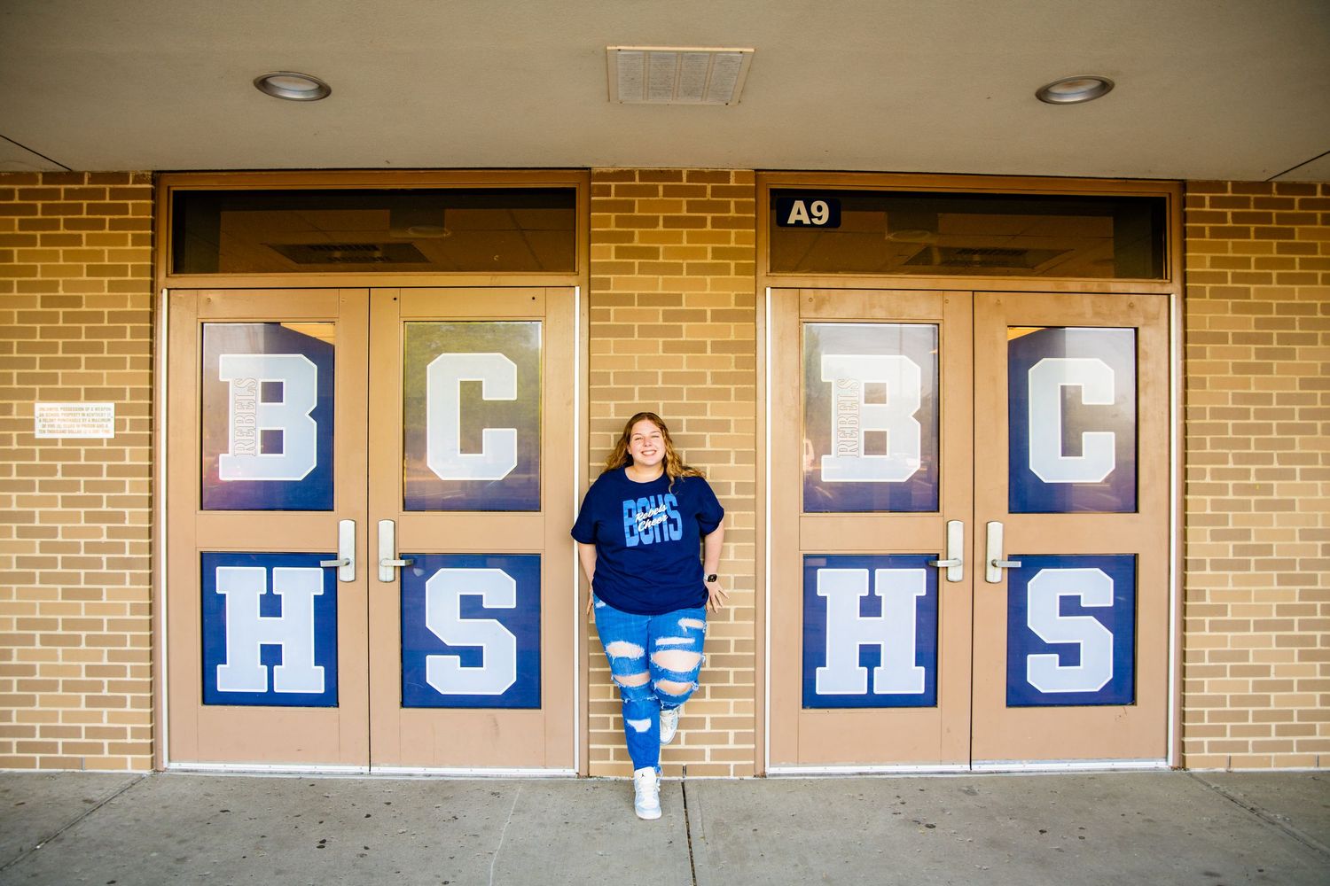 Student poses in front of BCHS school doors wearing blue shirt and tie-dye leggings at brick building entrance.