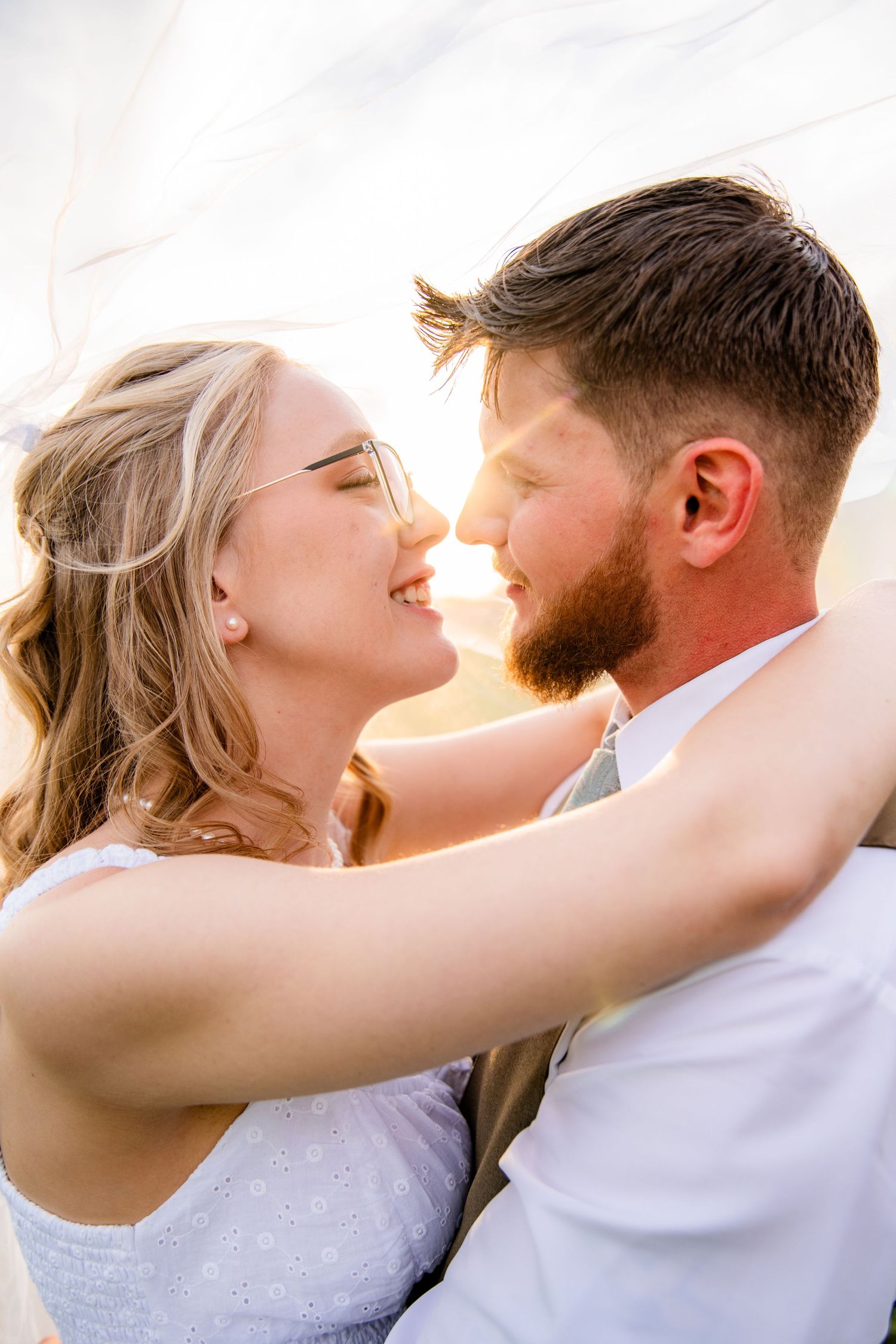 Bride and groom lean in for a kiss, laughing under flowing veil in warm golden sunlight.