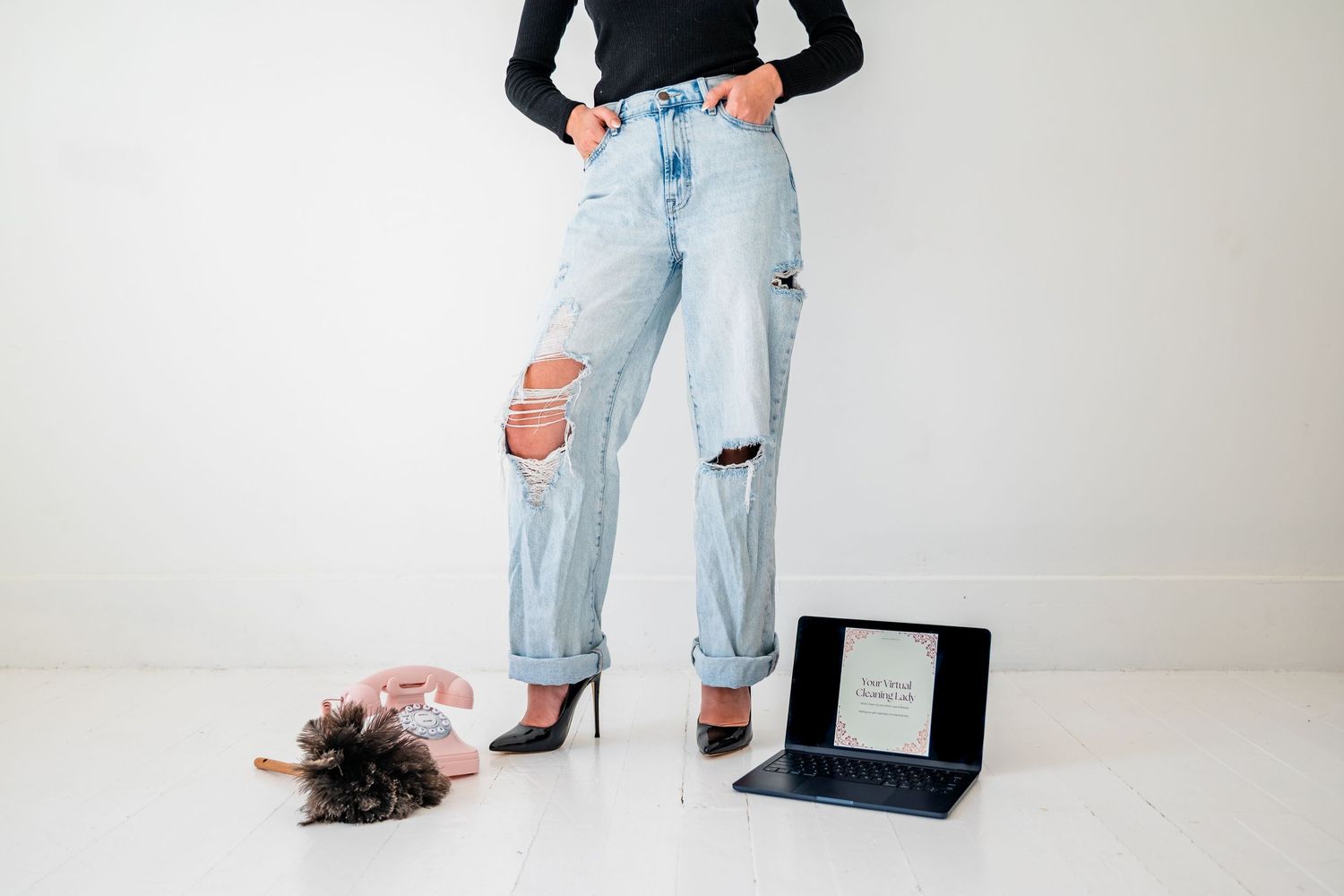 Woman in ripped light wash jeans and black heels standing next to a laptop and small dog on white floor.