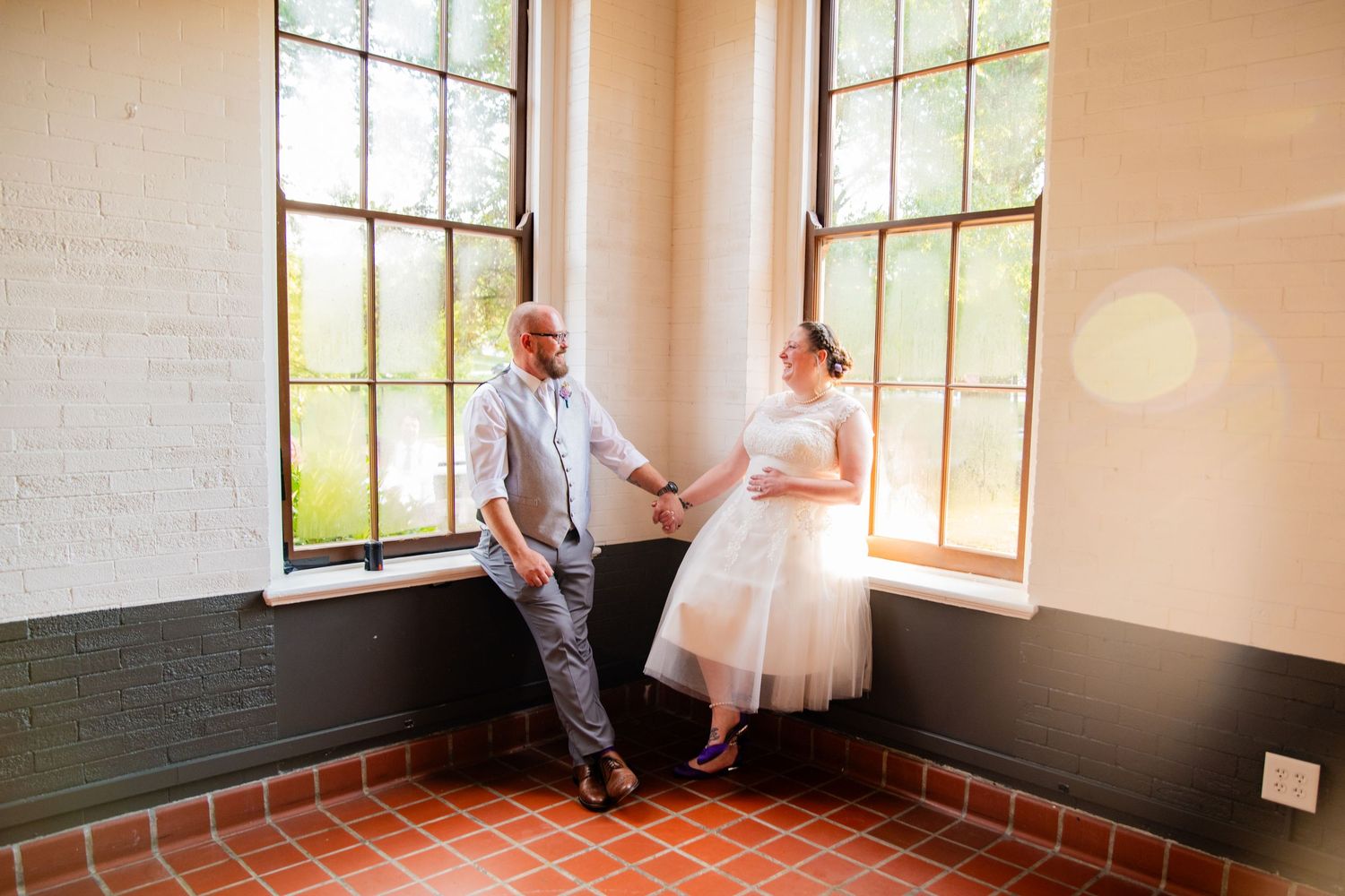 Couple holds hands and smiles in a sunlit room with large windows and terracotta tile floors during their wedding day.