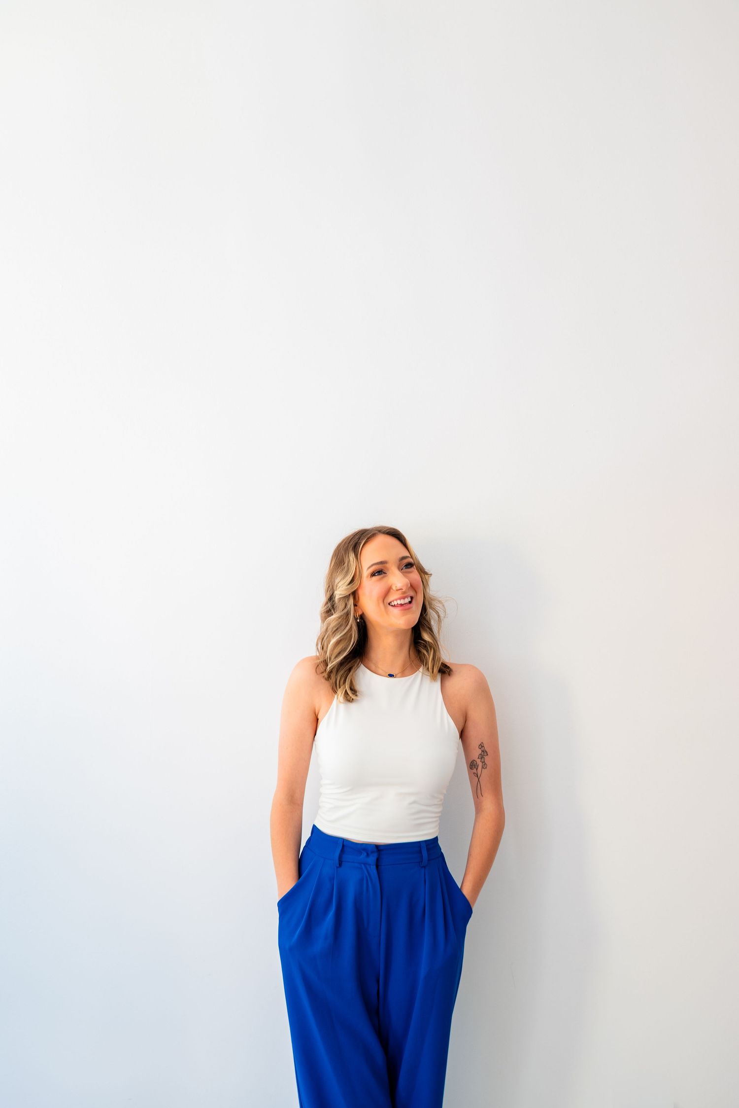 Smiling blonde woman in white sleeveless top and blue wide-leg trousers standing against a white wall.