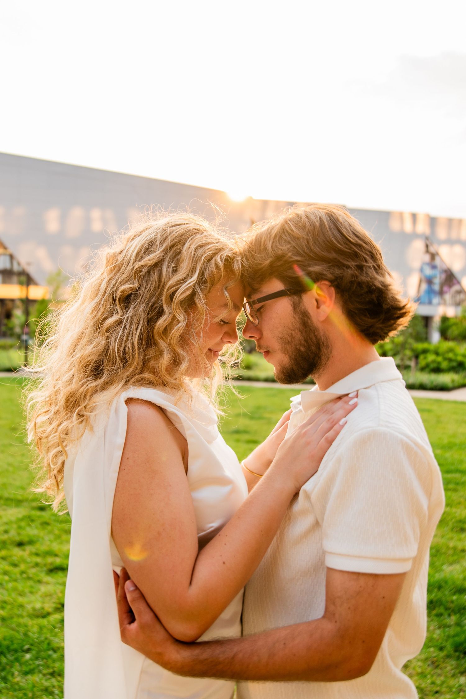 Romantic couple embracing outdoors at sunset, woman with curly blonde hair and man with beard wearing white outfits.