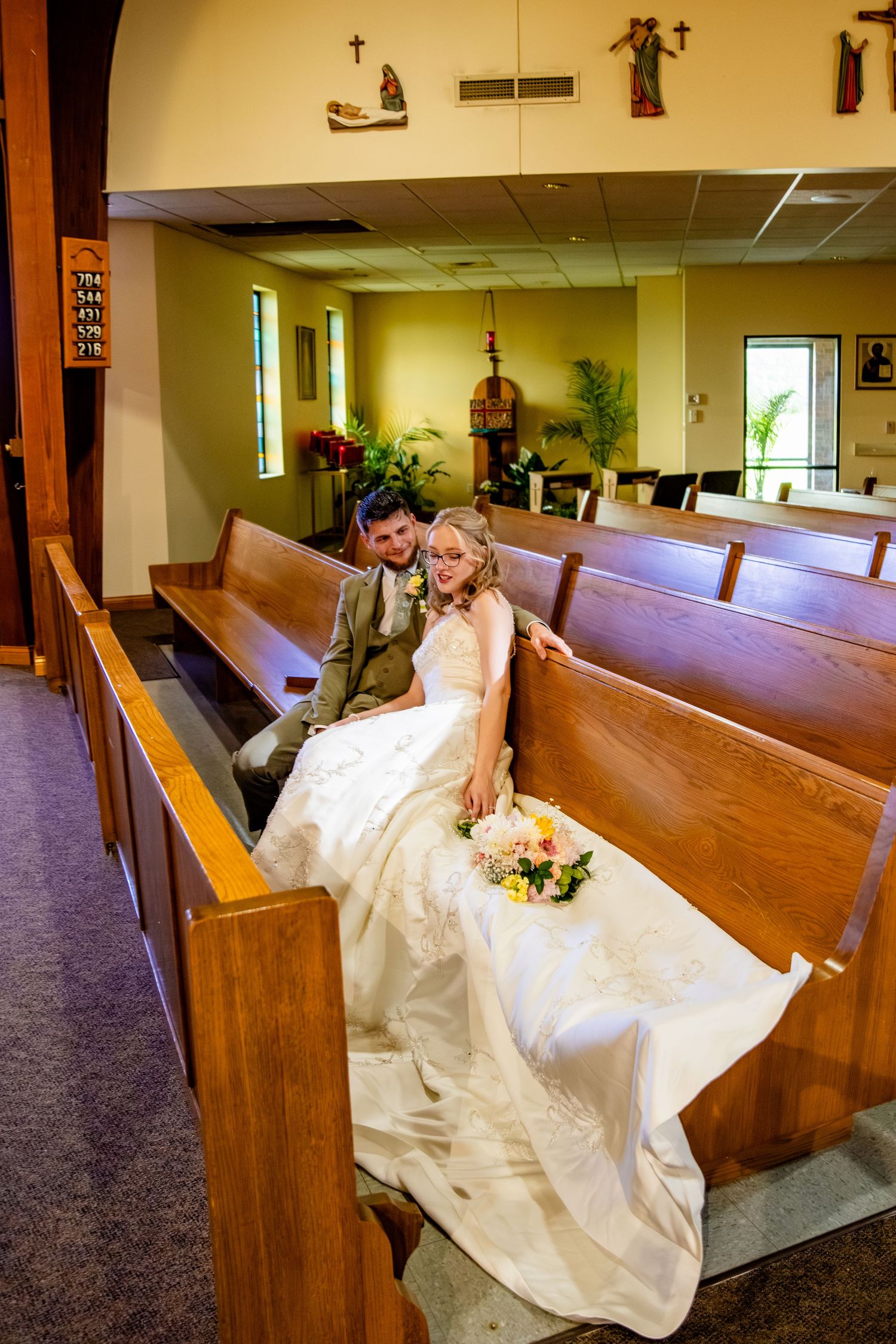 A bride and groom sit together on a wooden church pew with a bridal bouquet in a Catholic church.