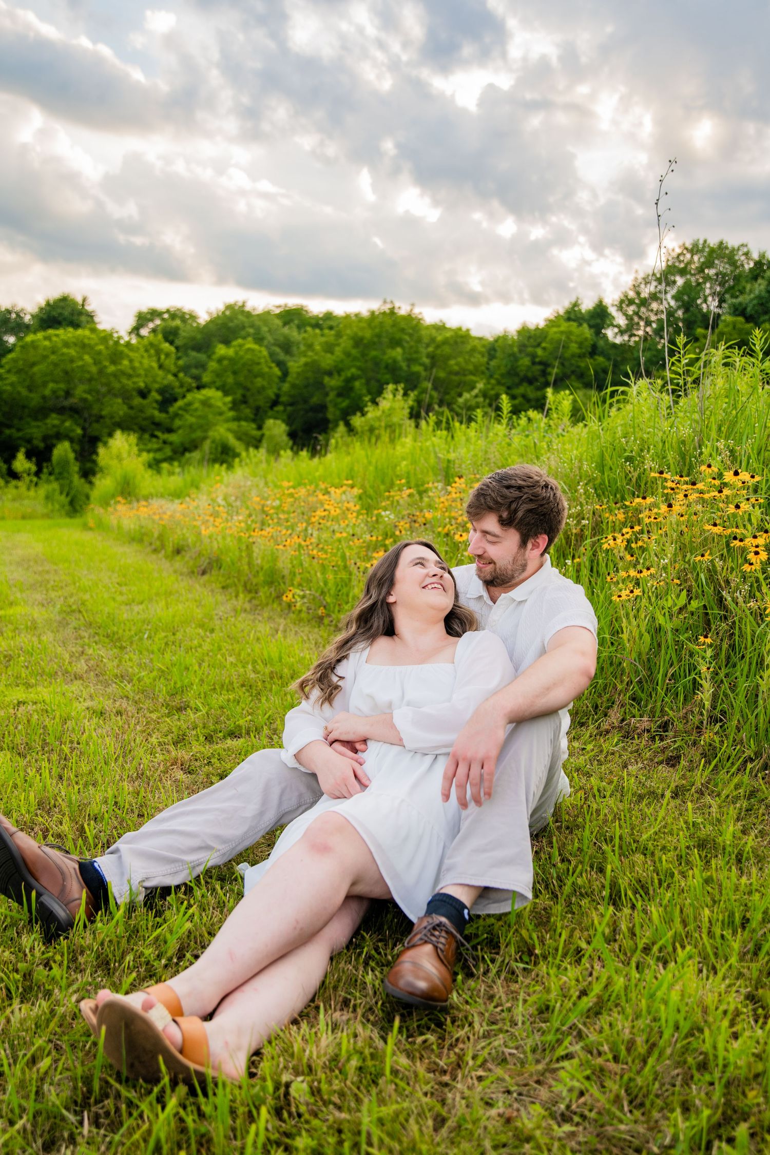 Couple sitting in a green meadow with wildflowers, sharing a romantic moment under a cloudy sky.