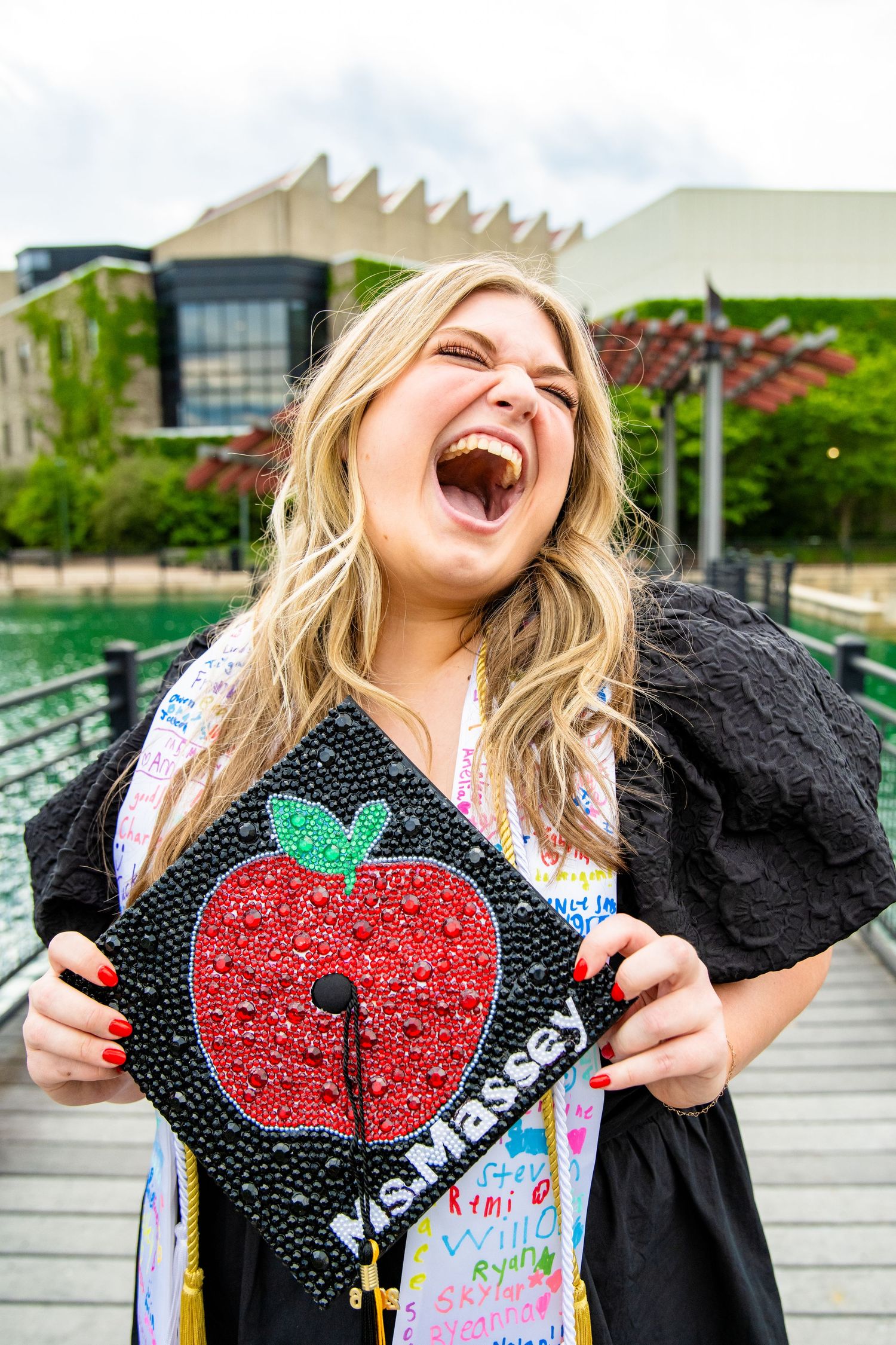Excited graduate holding decorated mortarboard cap with apple design and Ms. Massey name, laughing outdoors.