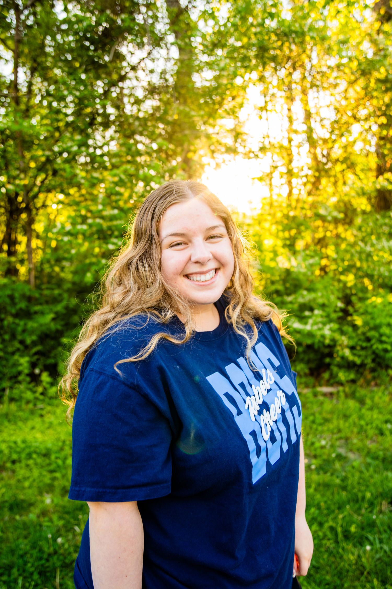Smiling young woman with blonde wavy hair wearing a navy blue t-shirt, standing outdoors in golden sunlight.