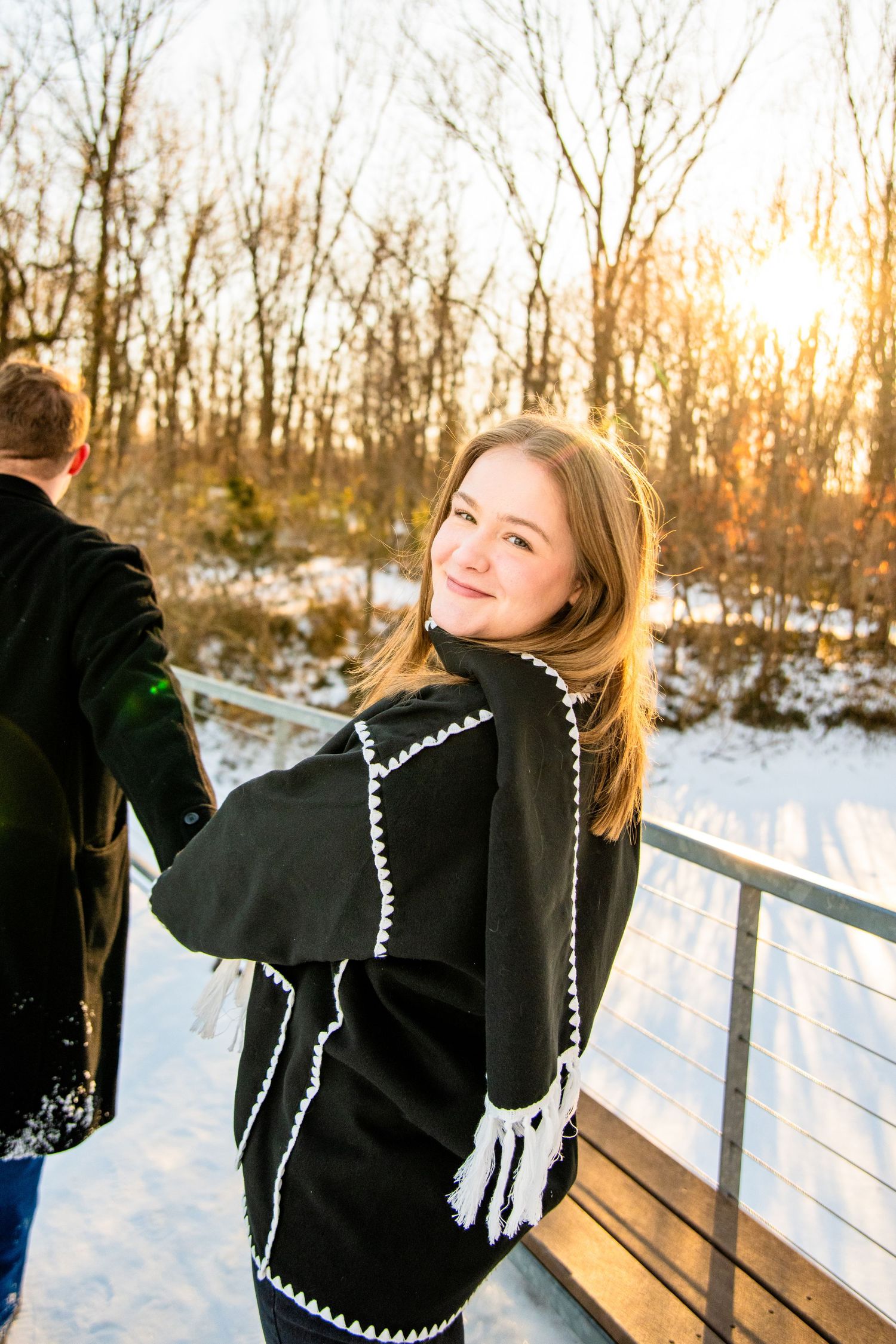 Young woman in black fringed shawl smiles over shoulder on snowy bridge at golden hour sunset.