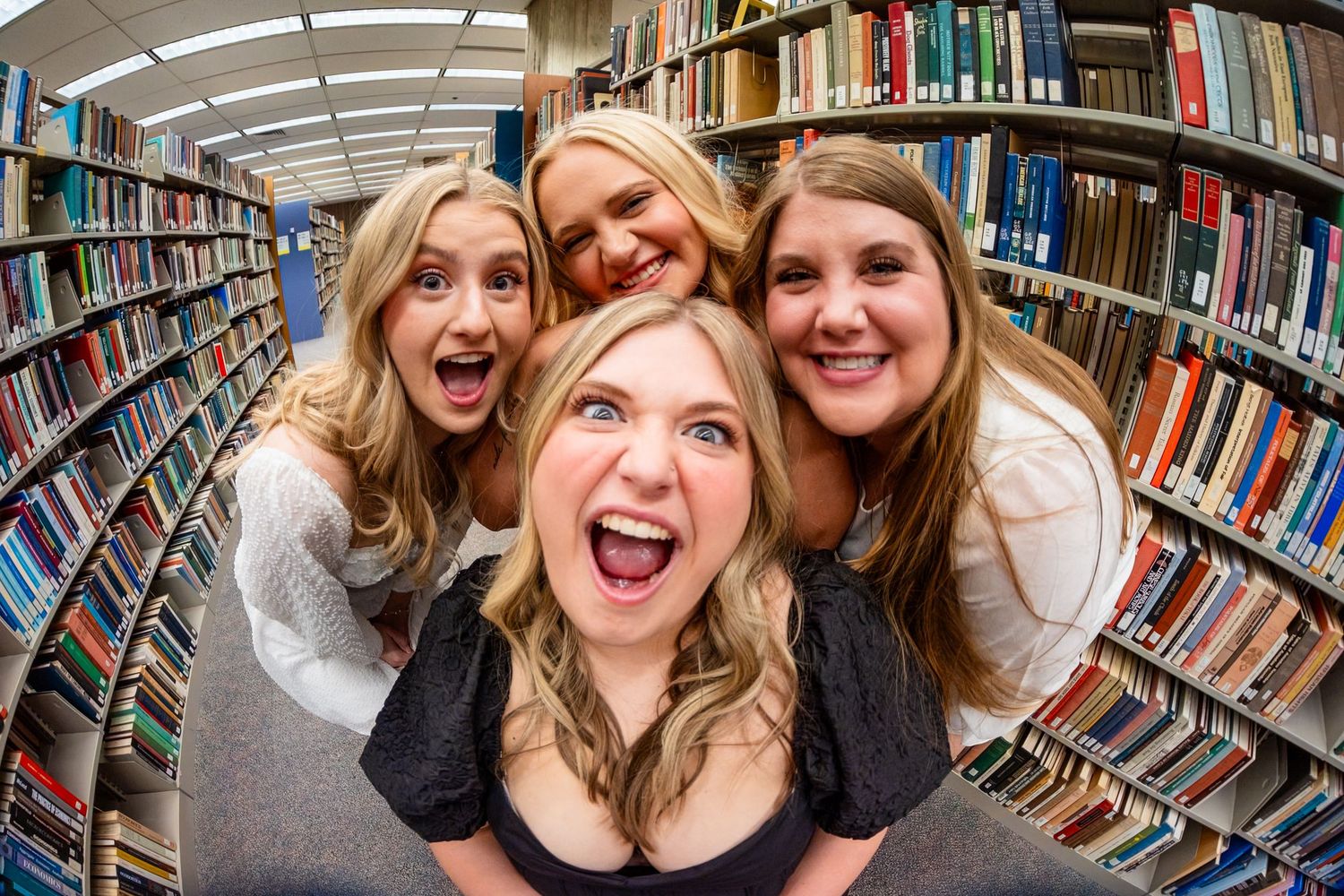 Four excited young women posing together in a library aisle, taking a fun fisheye lens selfie surrounded by bookshelves.