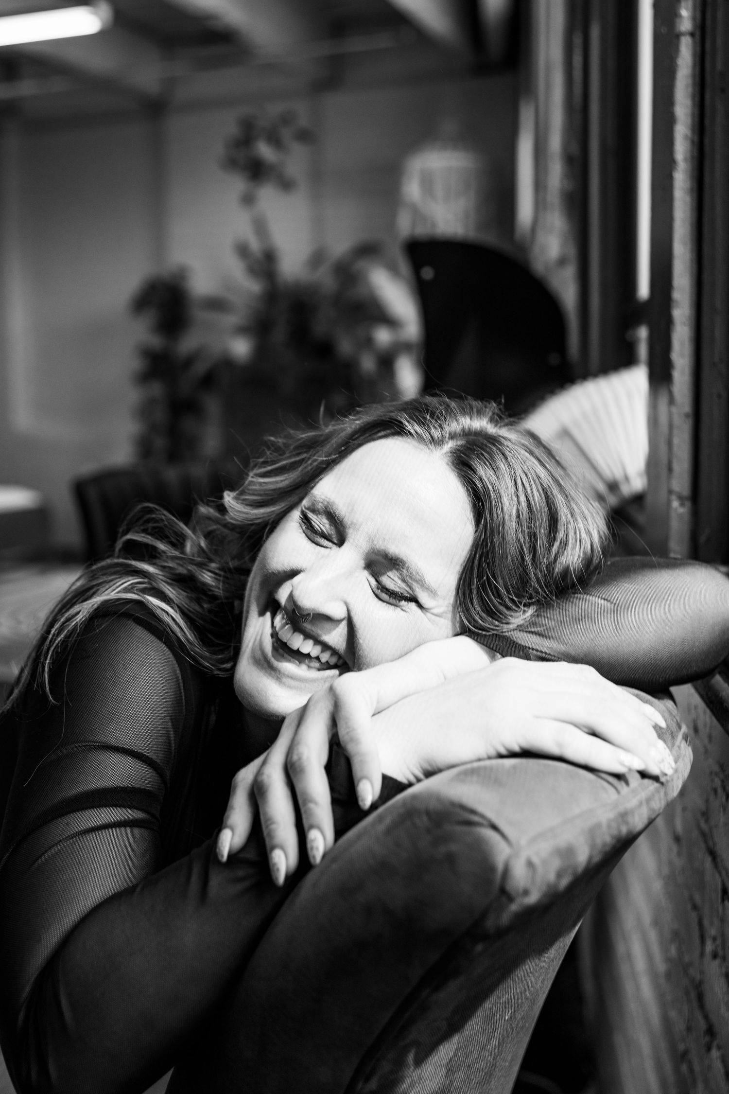 Black and white portrait of laughing woman resting chin on arms over sofa back in cozy indoor setting.