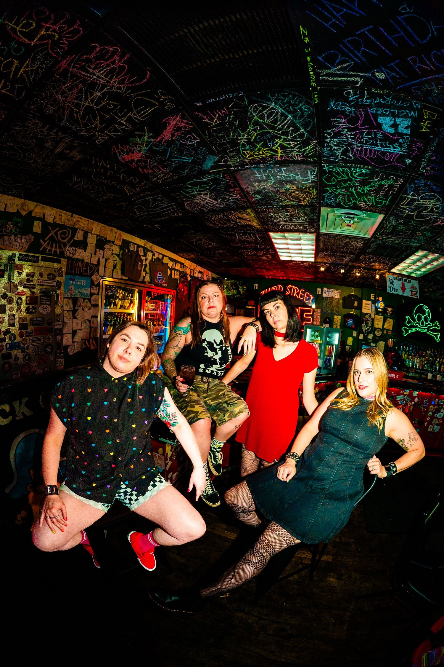 Four women pose energetically in a vibrant graffiti-covered bar with colorful neon lighting.