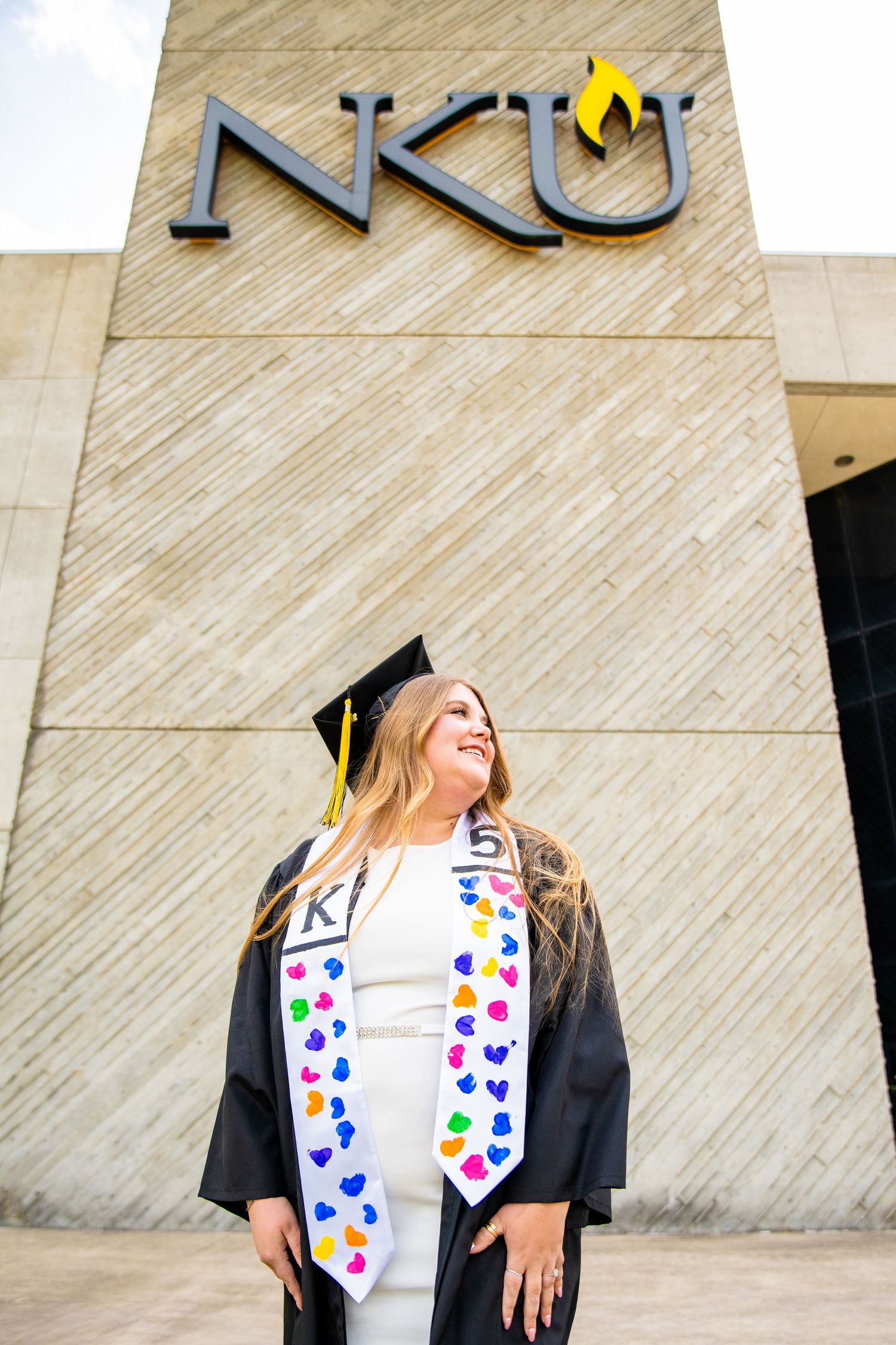 NKU graduate in cap and gown with colorful heart stole smiling proudly in front of Northern Kentucky University sign.