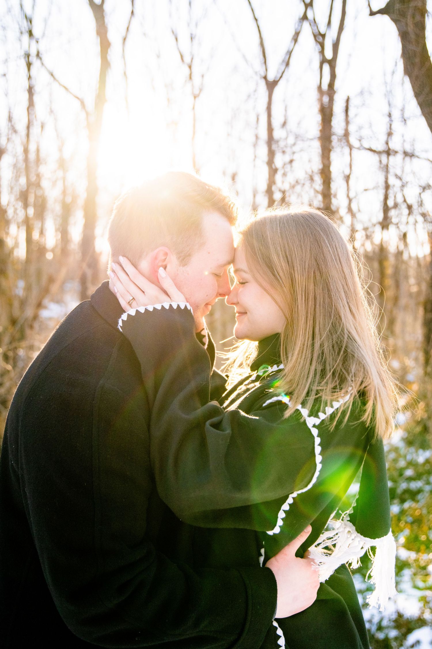 Couple sharing a romantic moment outdoors in winter, backlit by golden sunlight in a snowy forest setting.