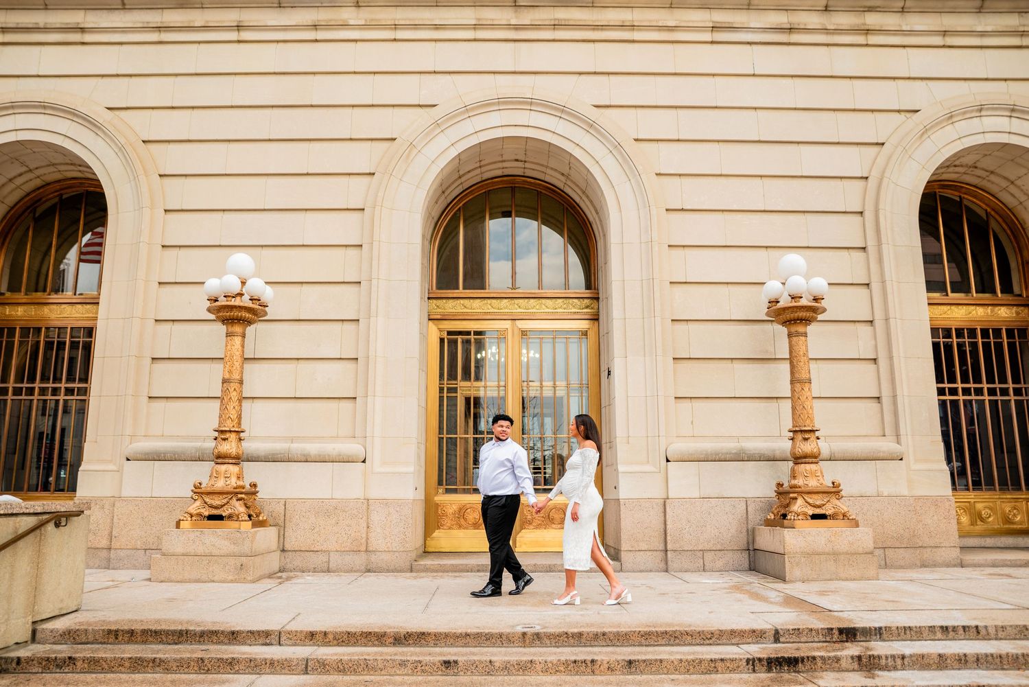 Couple in white attire walking on steps of grand beige stone building with ornate columns and arched windows.