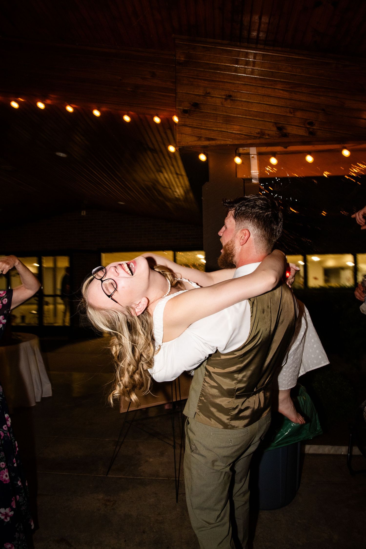 Bride laughing joyfully as groom dips her on the dance floor at their wedding reception under warm string lights.