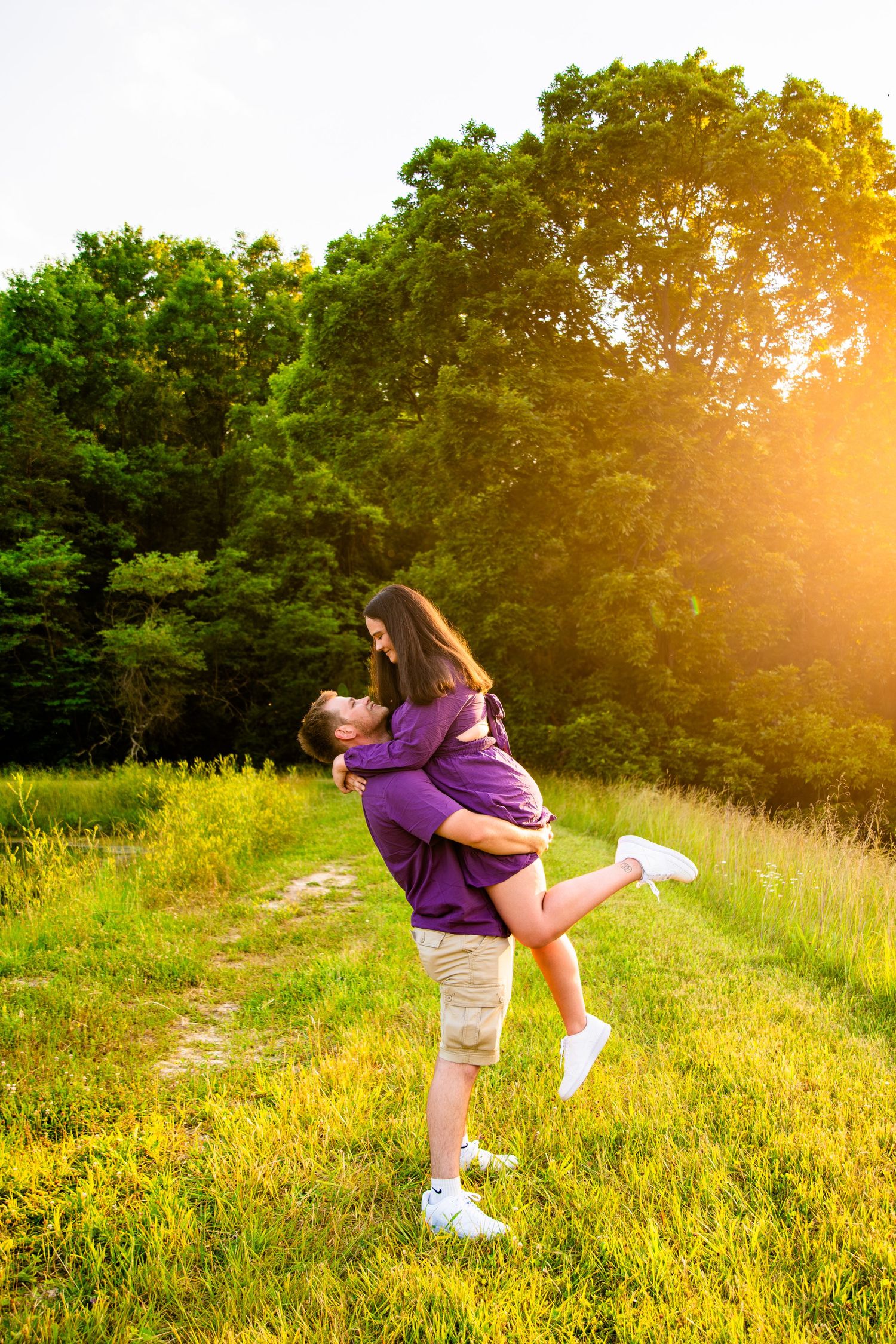 Couple in matching purple shirts embracing in a sunlit green meadow with trees in the background.