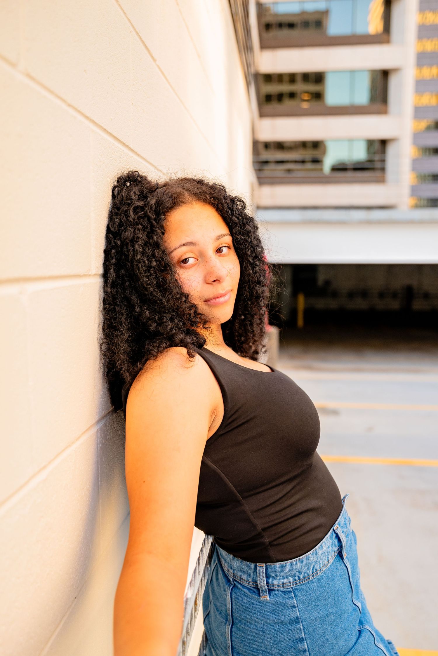 Young woman with curly hair leans against a wall in an urban setting, wearing a black tank top and jeans.
