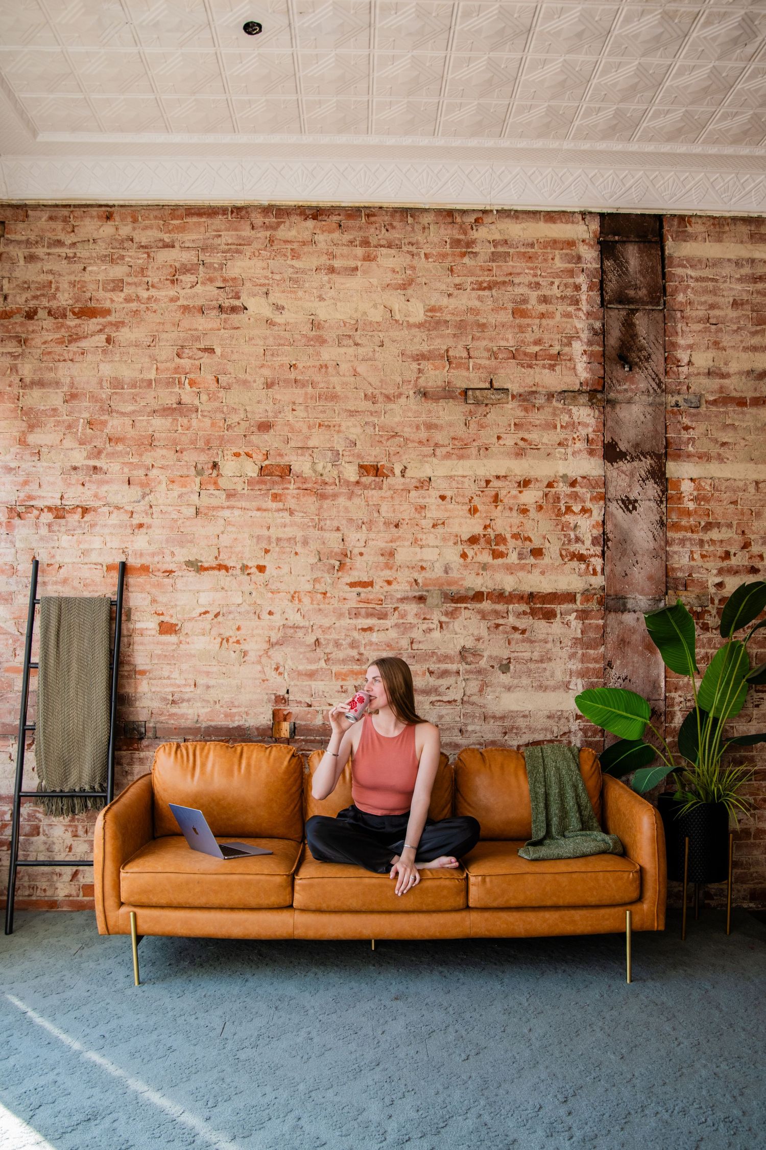 Woman sitting cross-legged on tan leather sofa with laptop, sipping drink near exposed brick wall and tropical plant.