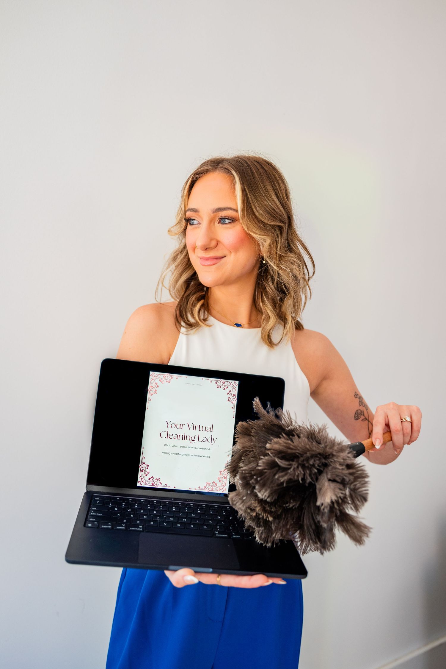Woman holding laptop displaying Your Virtual Cleaning Lady website while holding a feather duster, wearing blue skirt.
