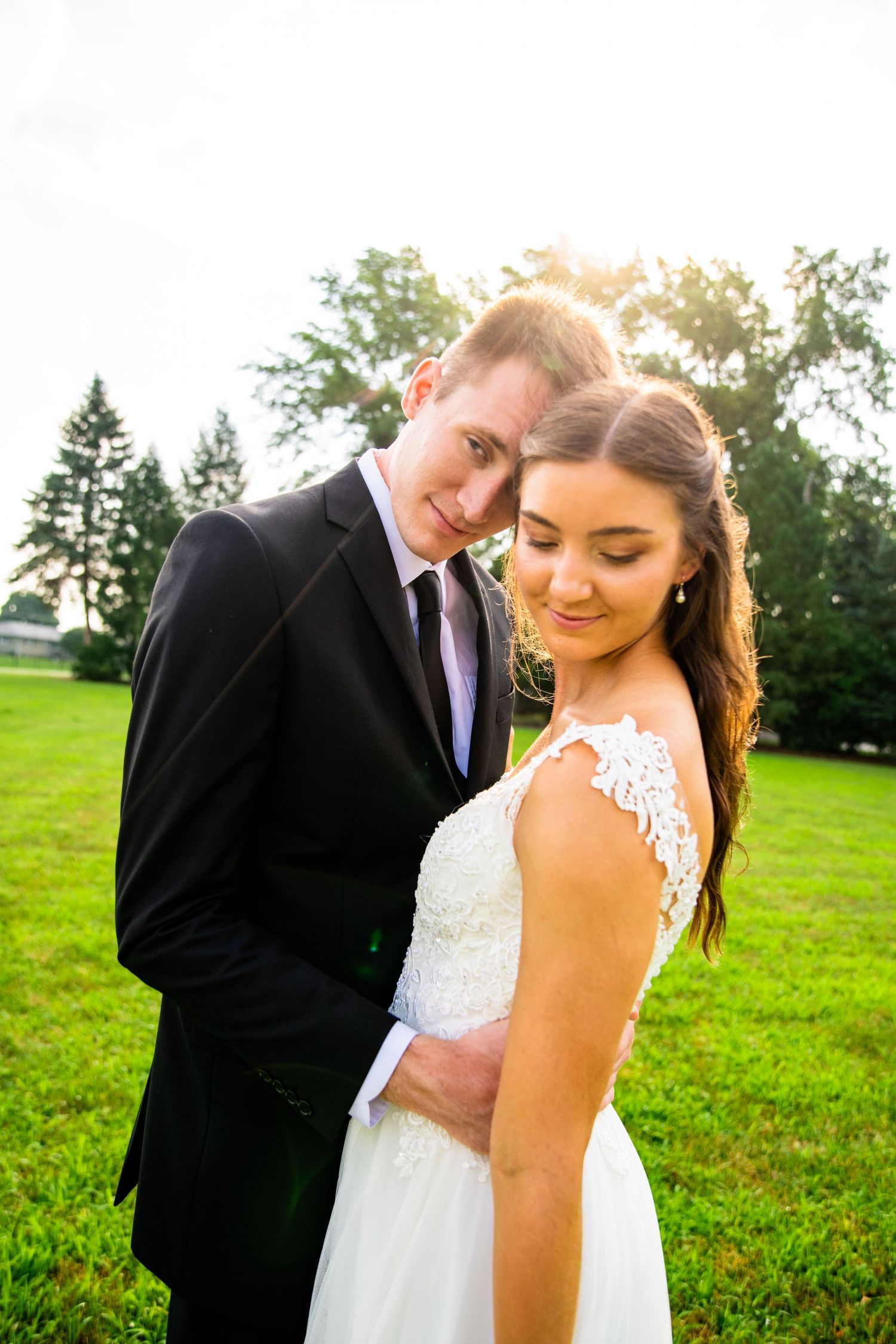 Groom in black suit tenderly leans toward bride in lace wedding dress in a sunlit green field.