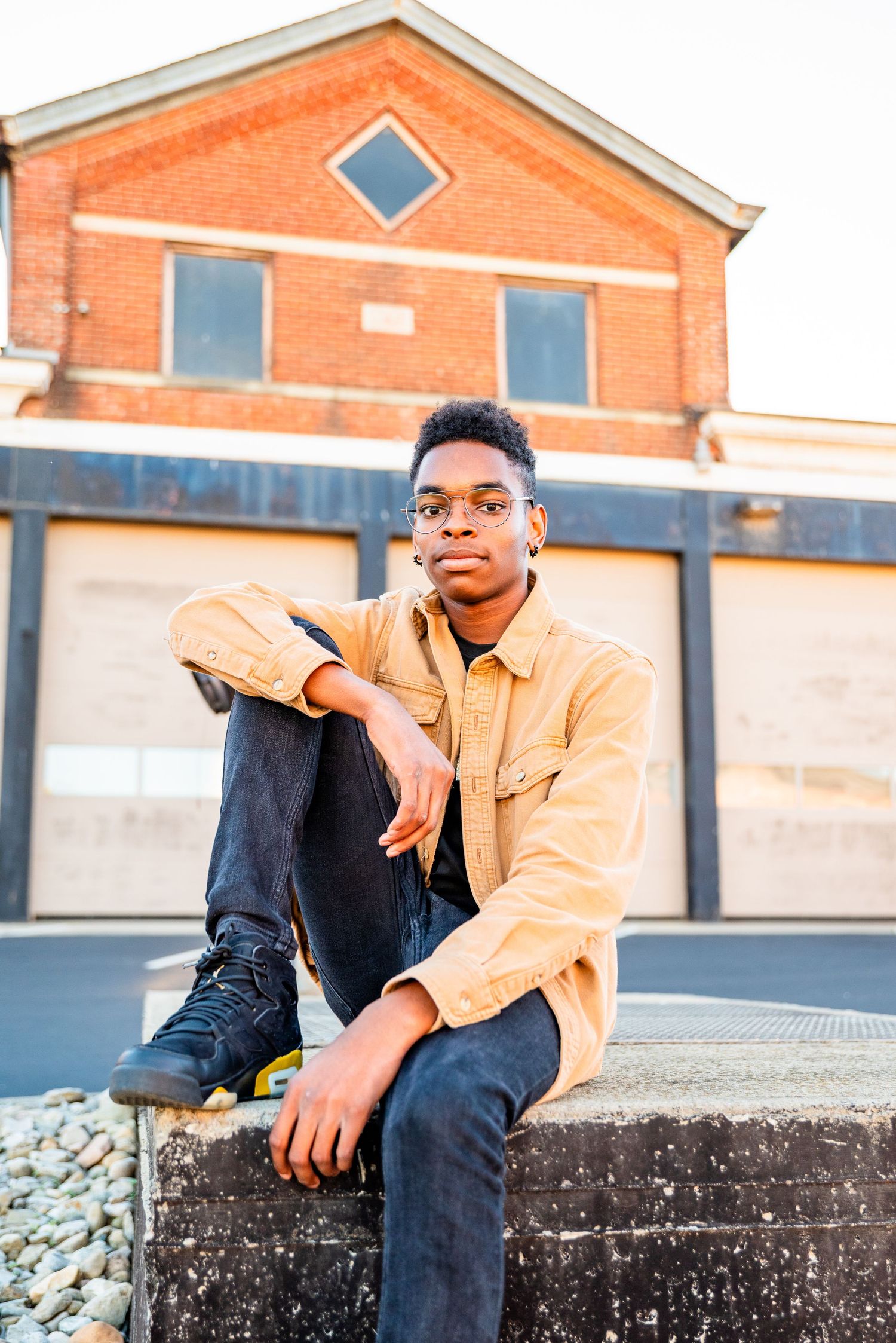 A young Black teen in a yellow jacket and jeans sits on a ledge outdoors near a brick building in warm sunlight.