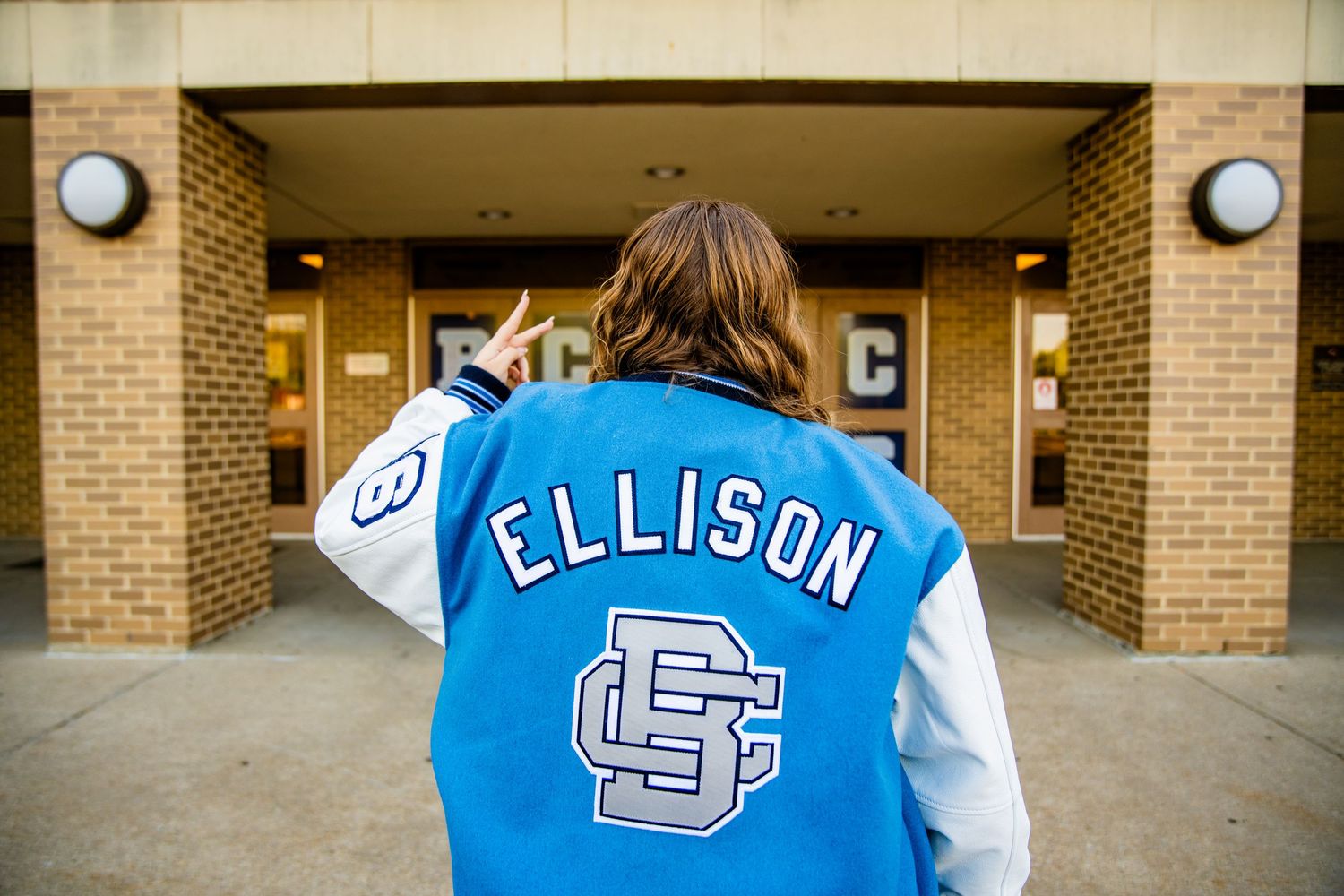 Person wearing a blue Ellison varsity letterman jacket standing in front of a brick school building.