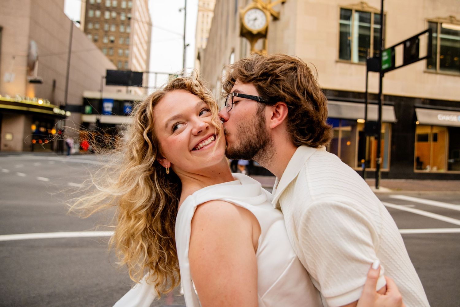 Smiling woman being kissed on the cheek by her partner on a busy city street corner.