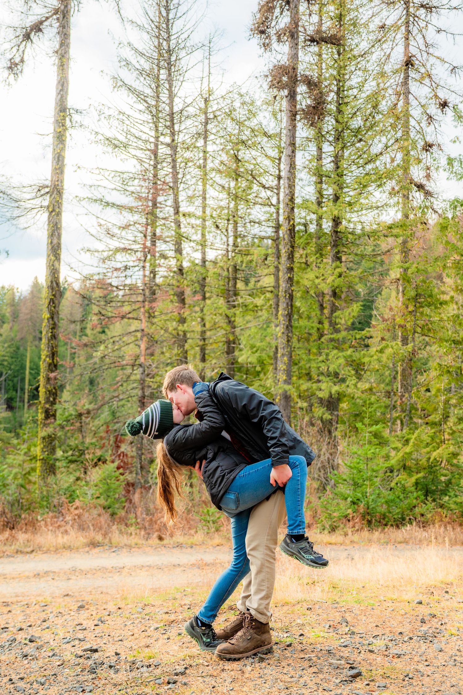 Couple playfully piggyback riding on a gravel path surrounded by tall evergreen trees in a forest.