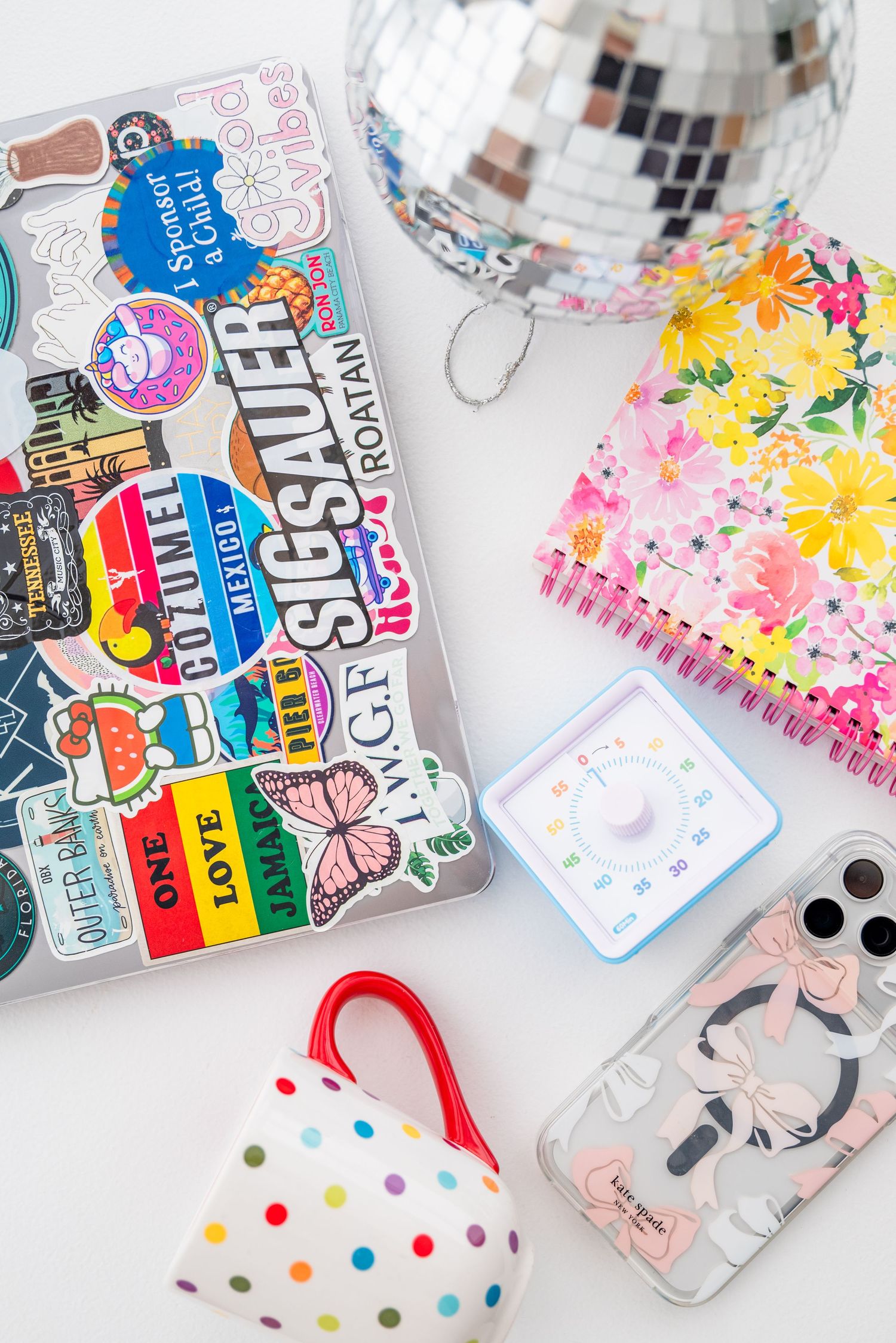 Colorful flatlay with sticker-covered laptop, disco ball, floral notebook, polka dot bag, phone case, and timer.