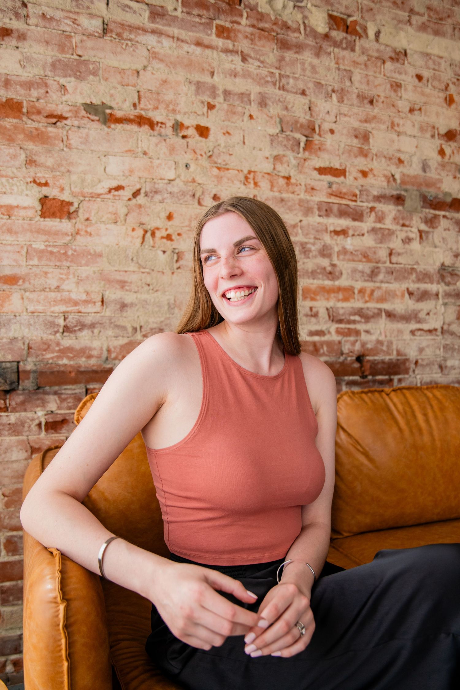 Smiling young woman in coral tank top sitting on brown leather sofa against rustic brick wall.