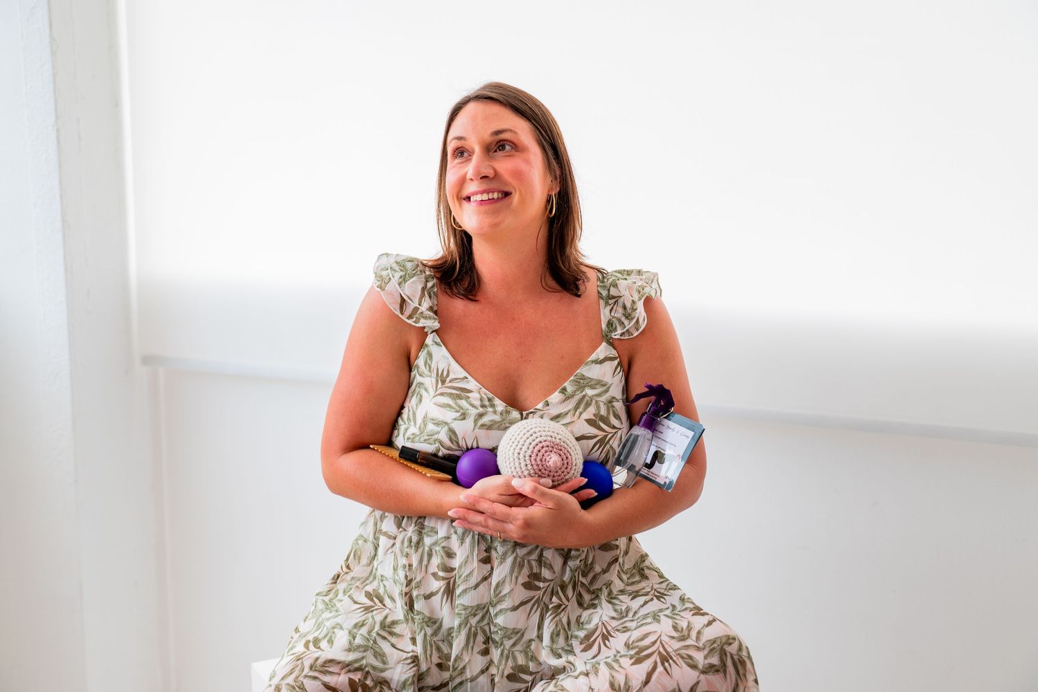 Woman in floral dress smiling while holding beauty and wellness products in a bright white room.