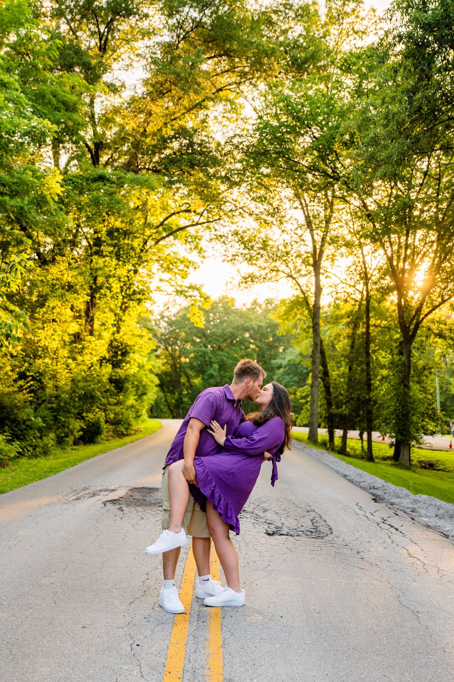 Couple sharing a romantic kiss on a tree-lined road at golden sunset, woman in purple dress lifted by partner.