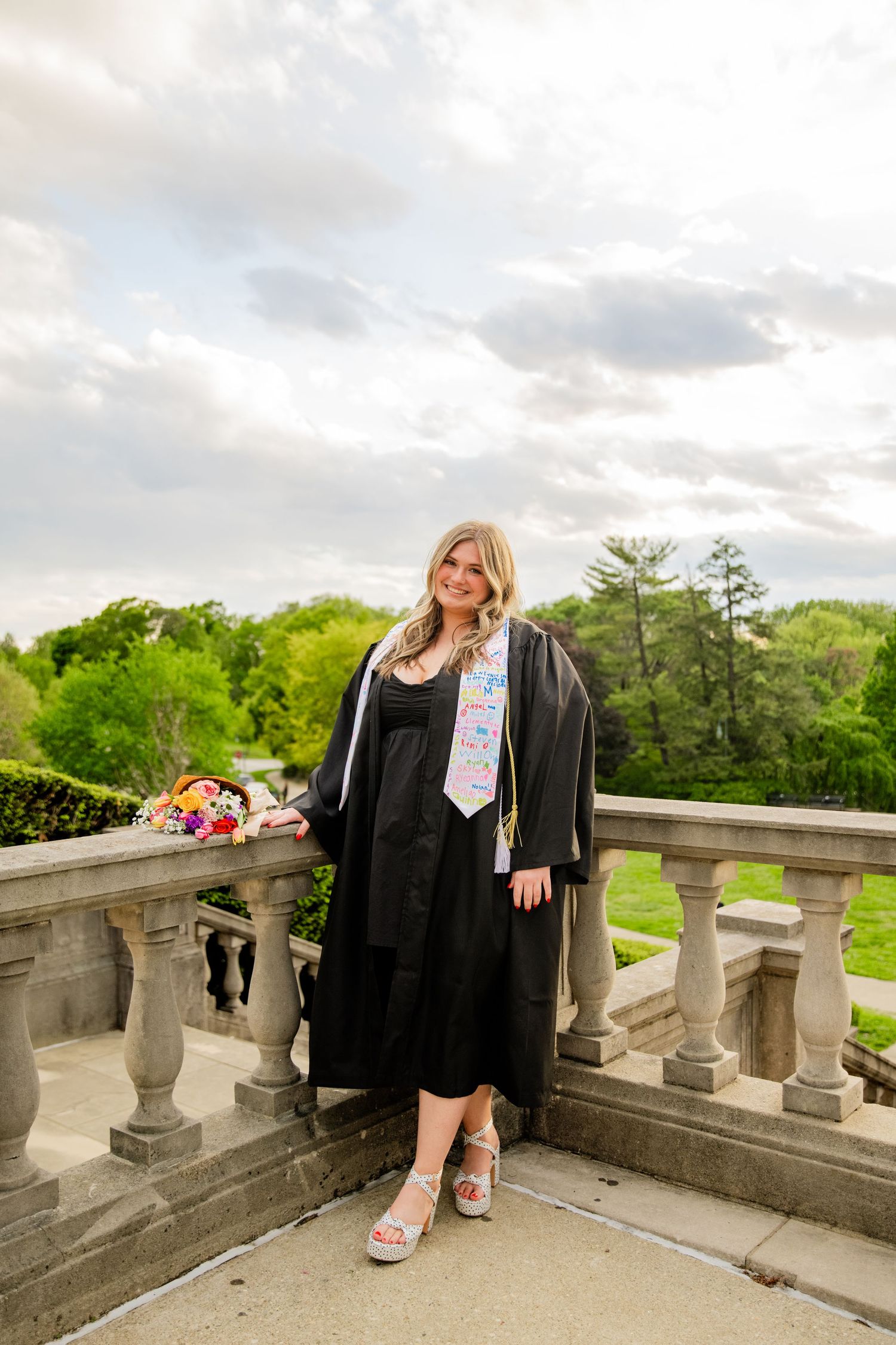 A smiling graduate in black cap and gown poses on a stone balcony surrounded by lush greenery.