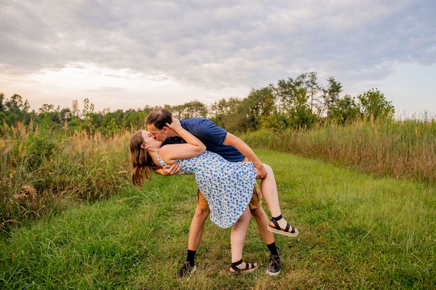 Couple sharing a romantic dip kiss in a lush green meadow field at sunset during an outdoor engagement photo session.