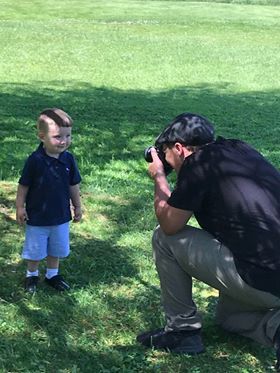 Man crouching to photograph a smiling toddler boy in navy polo and shorts on a sunny green lawn.