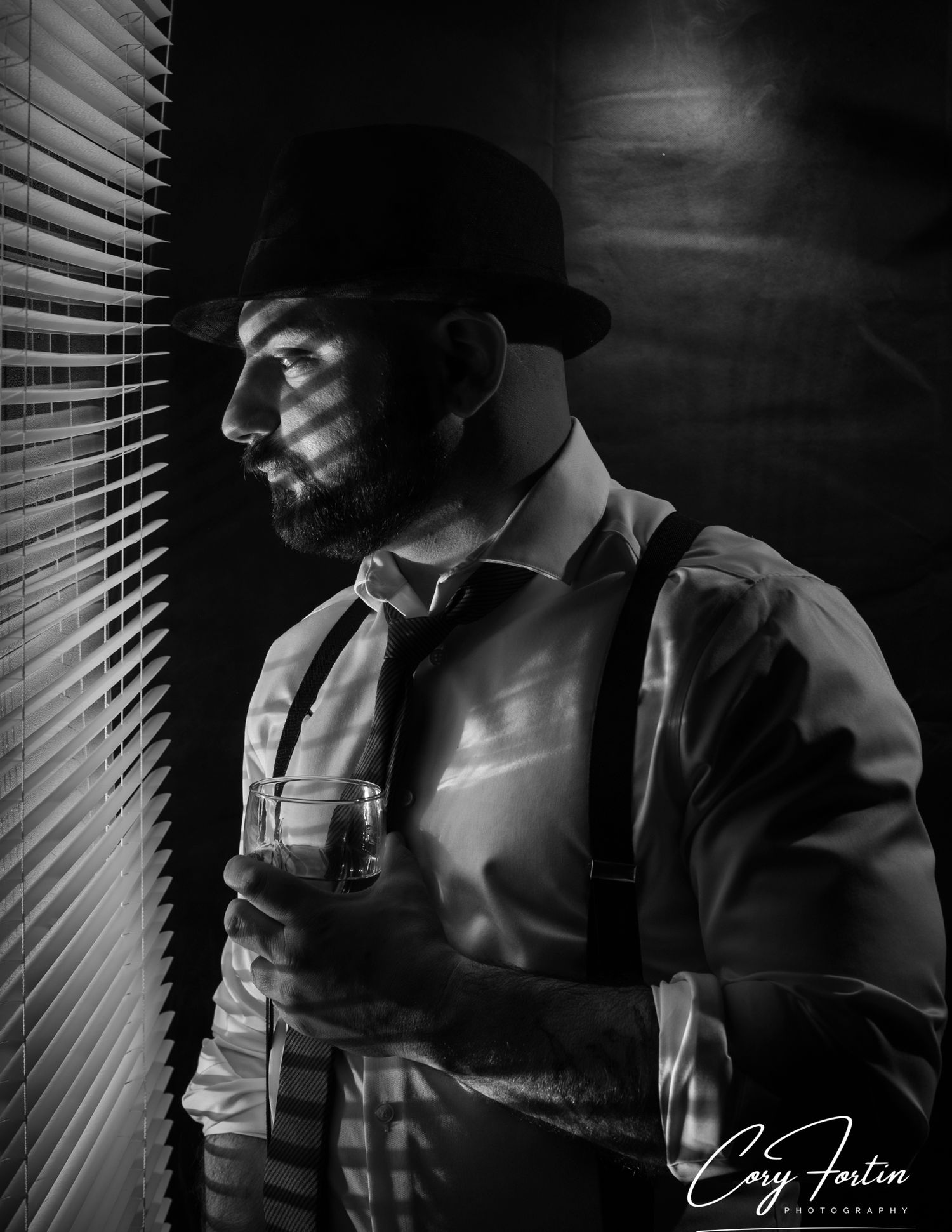 Black and white portrait of a bearded man in suspenders and bowler hat holding a glass near window blinds.