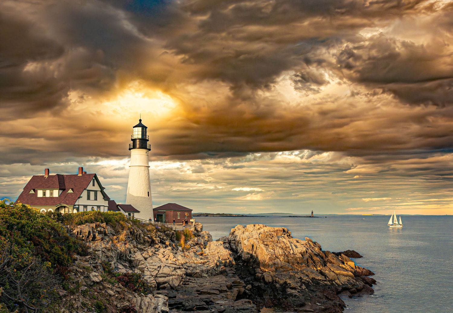 Dramatic stormy sunset sky over Portland Head lighthouse on rocky Maine coast with sailboat on calm ocean waters.