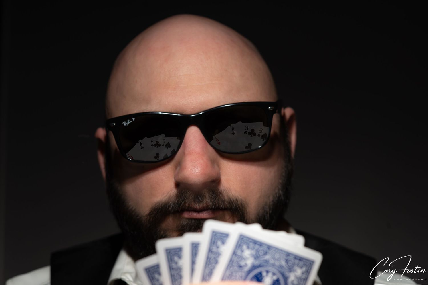 Bald man with beard and sunglasses holding a fan of playing cards in dramatic studio lighting.