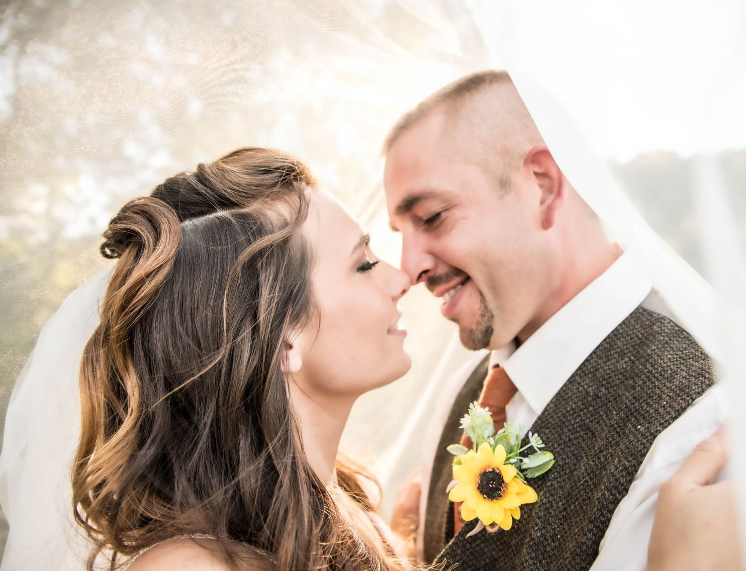 Bride and groom share a romantic almost-kiss under a veil, groom wearing a sunflower boutonniere on his tweed vest.