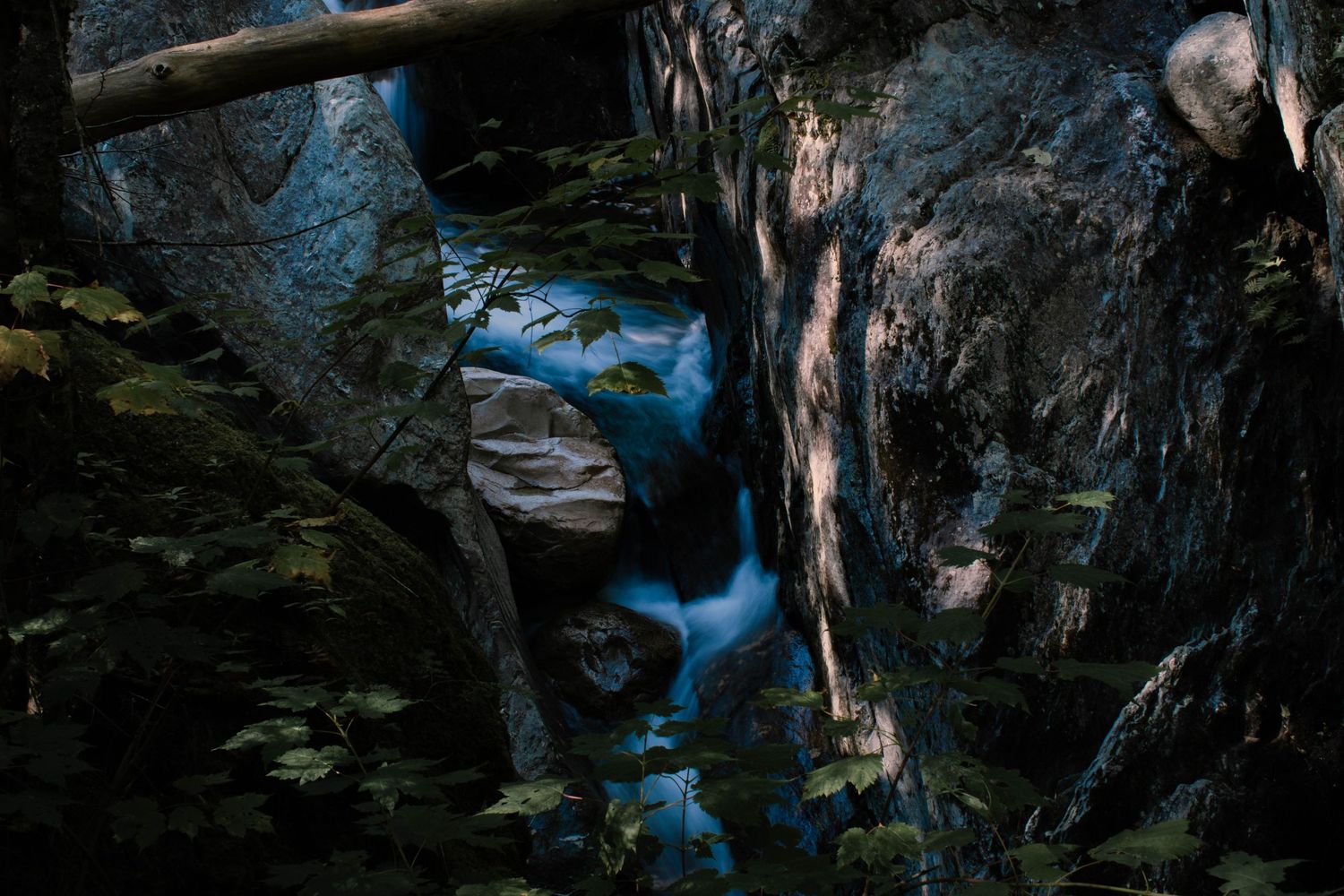 A serene blue stream flows through dark rocky canyon walls surrounded by lush green foliage in a forest.