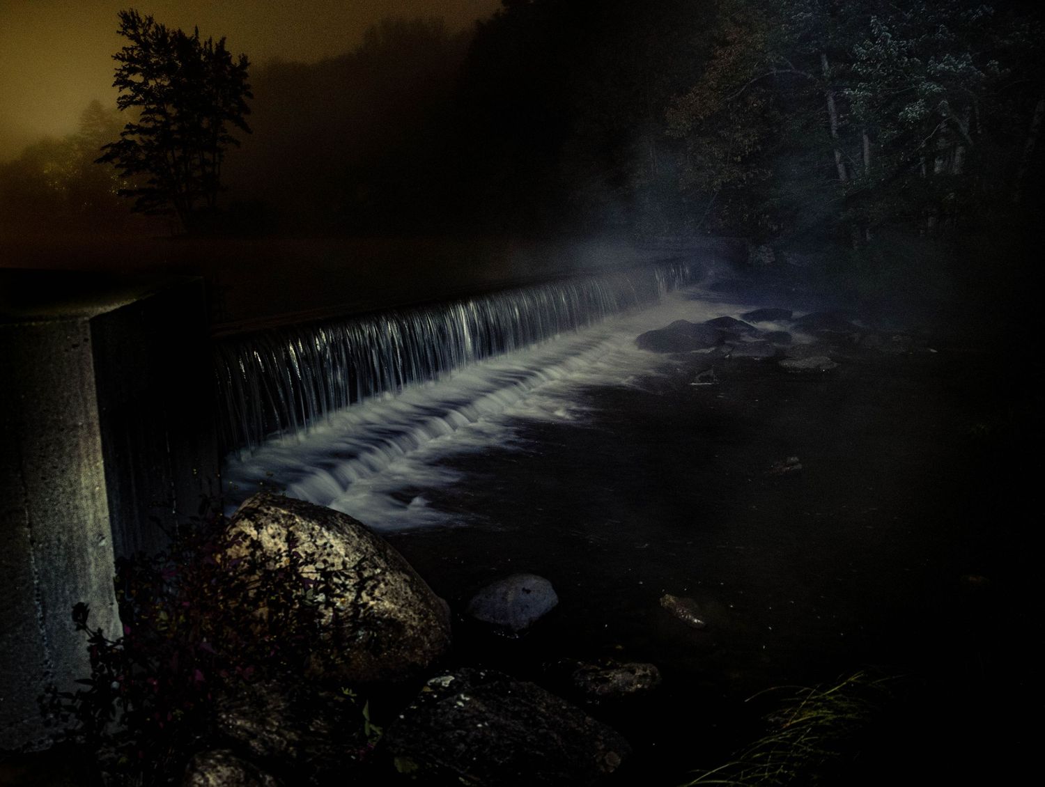 A misty waterfall cascades over a dark stone dam at night, illuminated by soft light with trees in the foggy background.