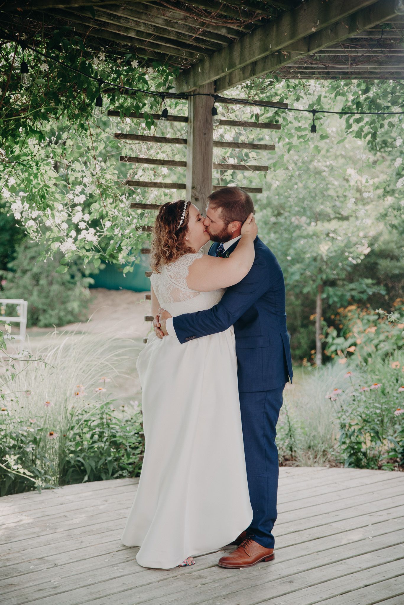Bride and groom share their first kiss under a wooden pergola surrounded by lush greenery.