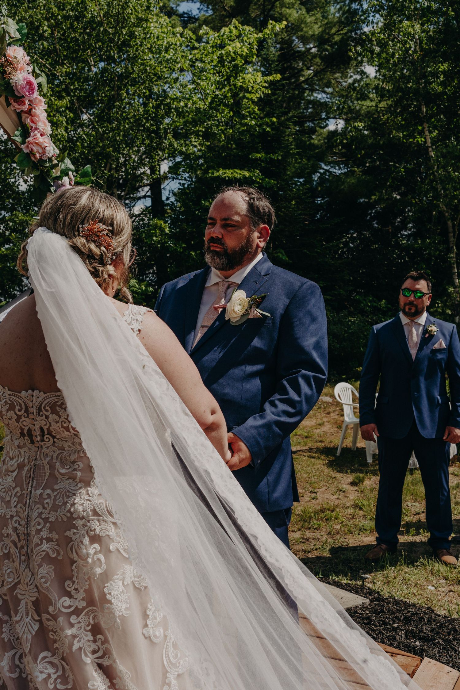 Groom in navy suit holding bride's hands during outdoor wedding ceremony in wooded setting.