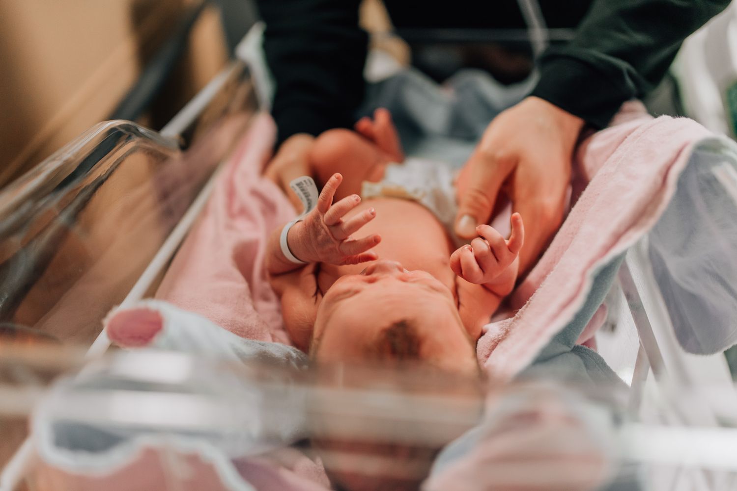 Newborn baby lying in hospital bassinet with adult hands gently touching the infant wrapped in pink blankets.