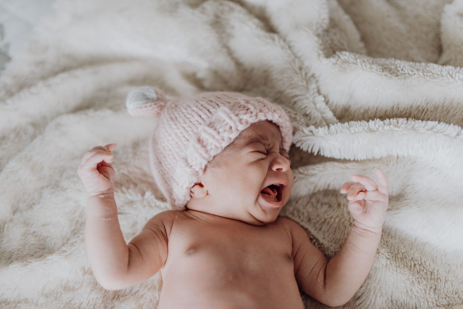 A newborn baby yawning and stretching on a soft white blanket, wearing a pink knit hat.